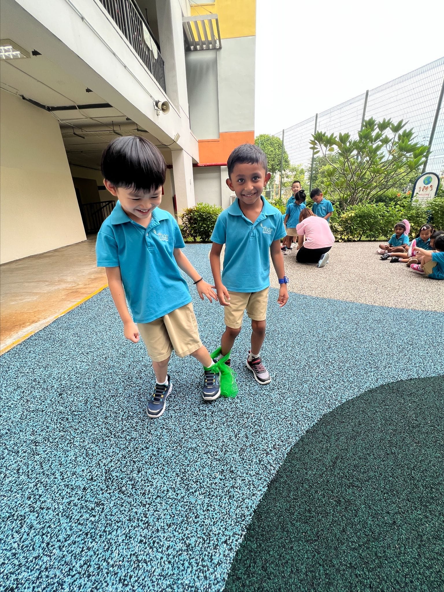 Two young boys in matching blue polo shirts and khaki shorts walk smiling on a colorful rubber playground surface.