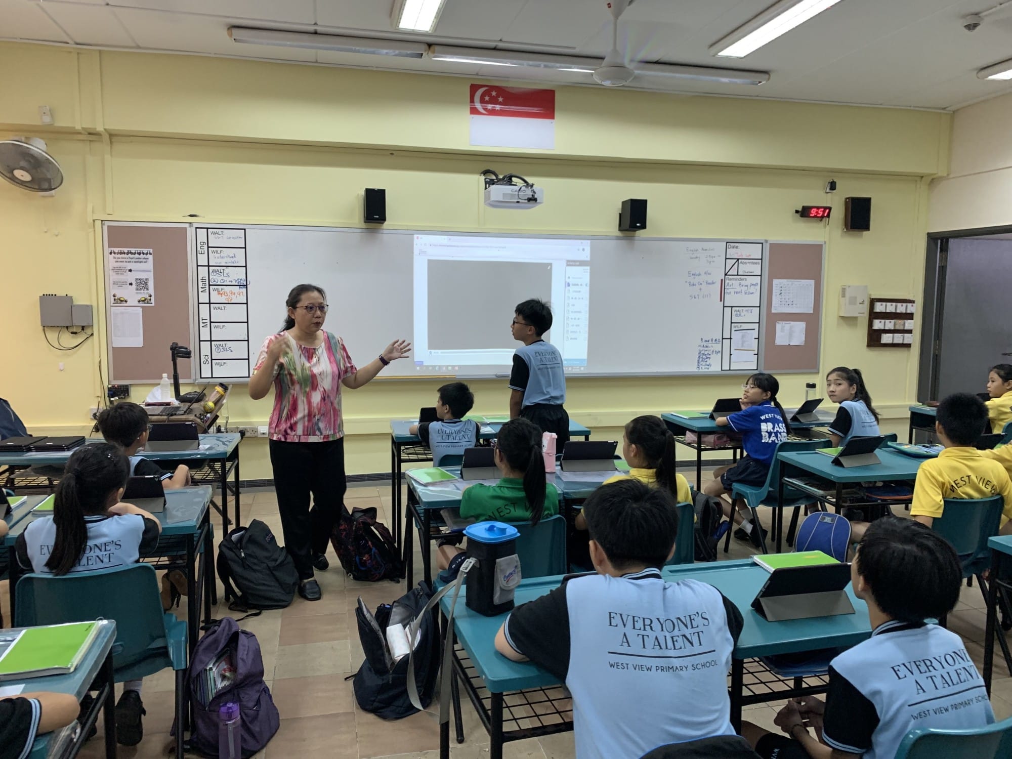 A teacher stands at the front of a classroom at West View Primary School, addressing seated students with a projected screen behind her.