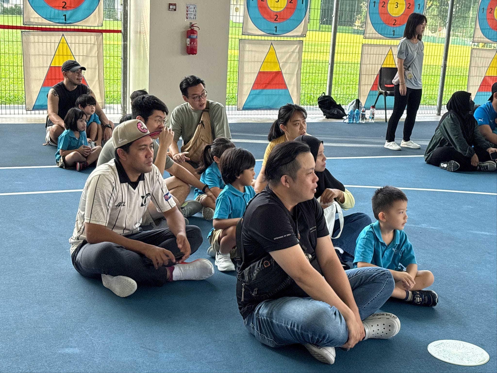 Adults and children in blue shirts sit on a blue floor in an indoor archery range with colorful targets on the walls.