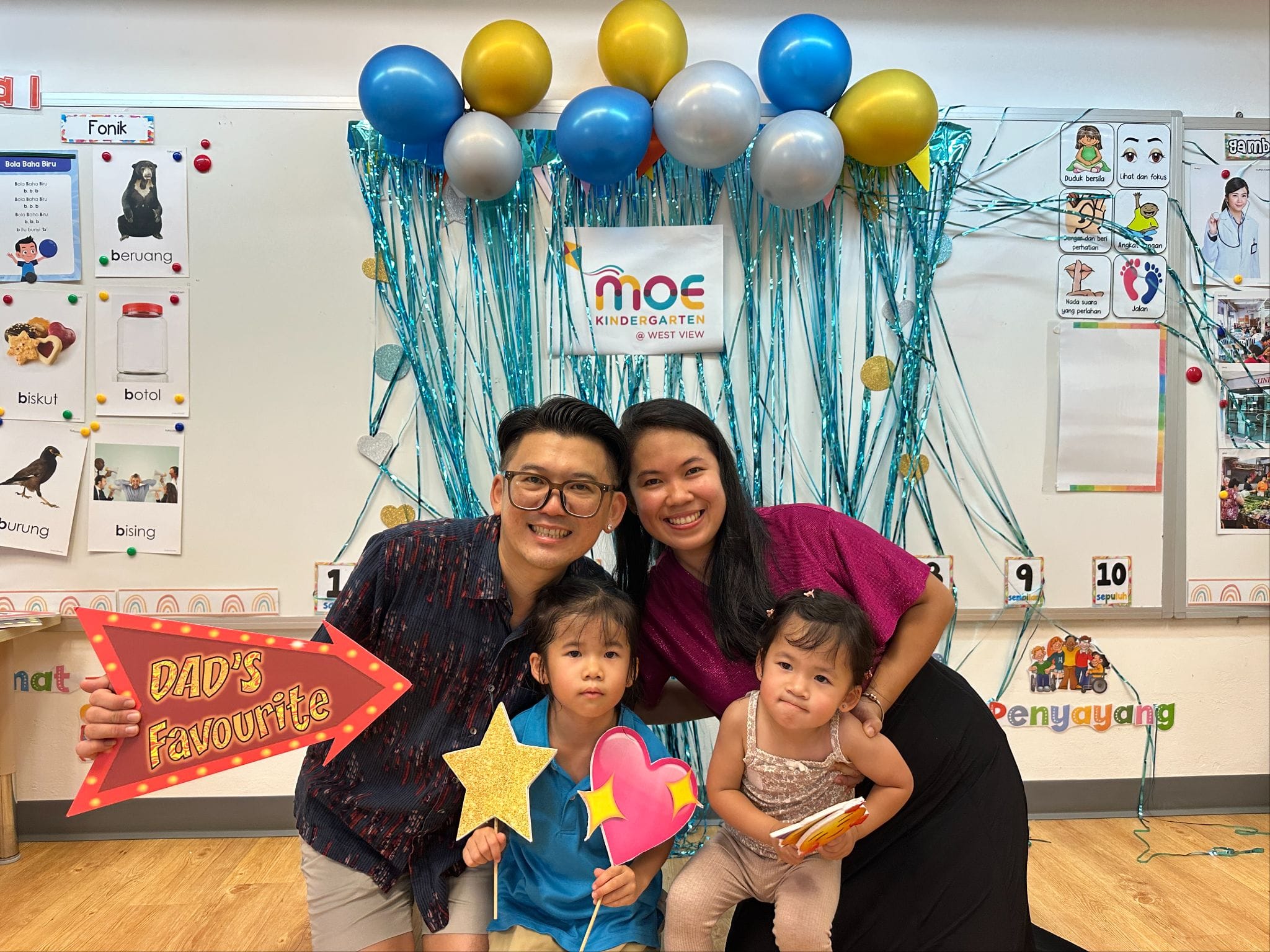 Family of four posing in front of a decorated MOE Kindergarten backdrop with blue, gold, and silver balloons.
