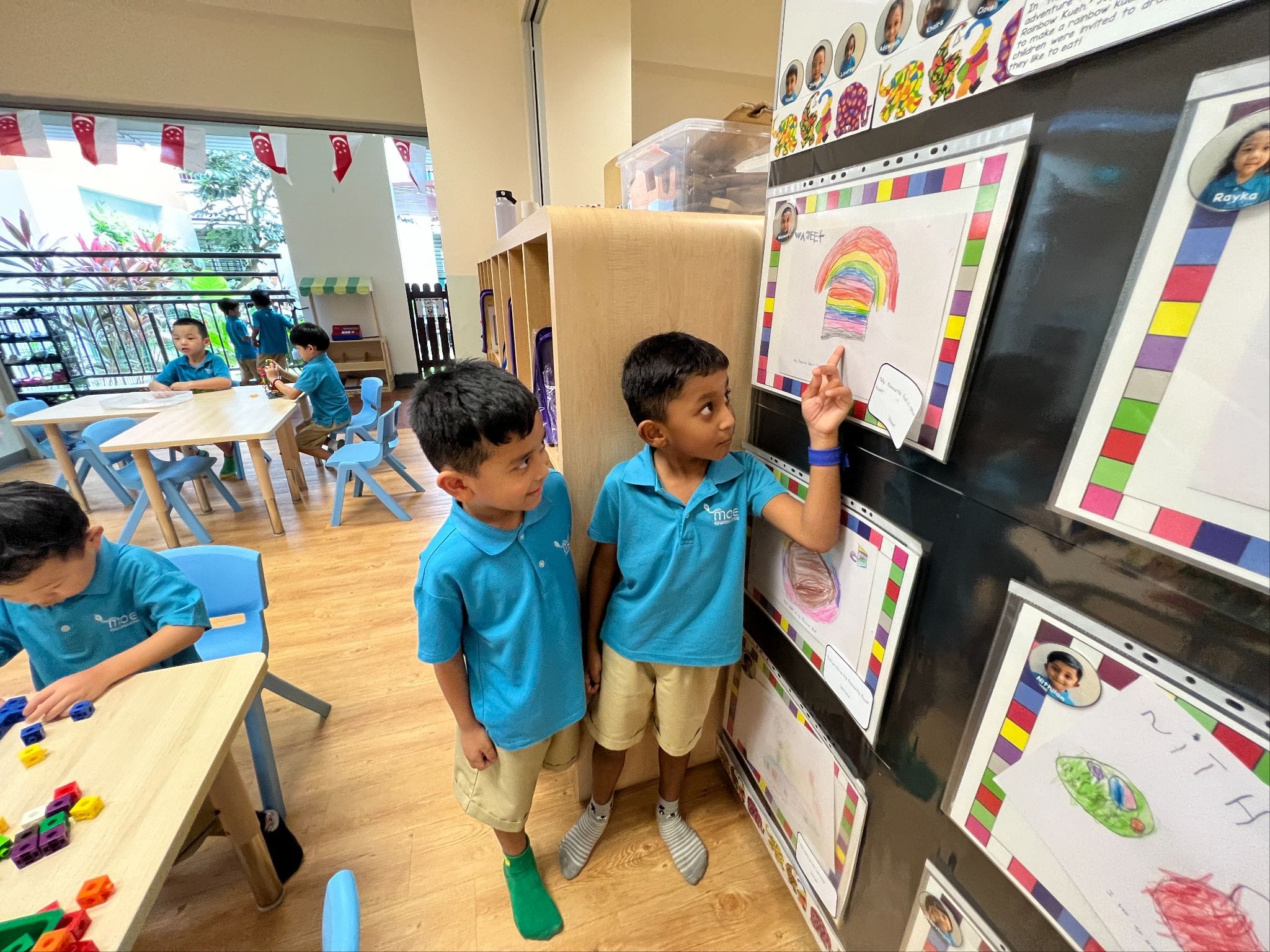 Two young boys in blue uniforms point at colorful children's artwork displayed on a classroom wall.