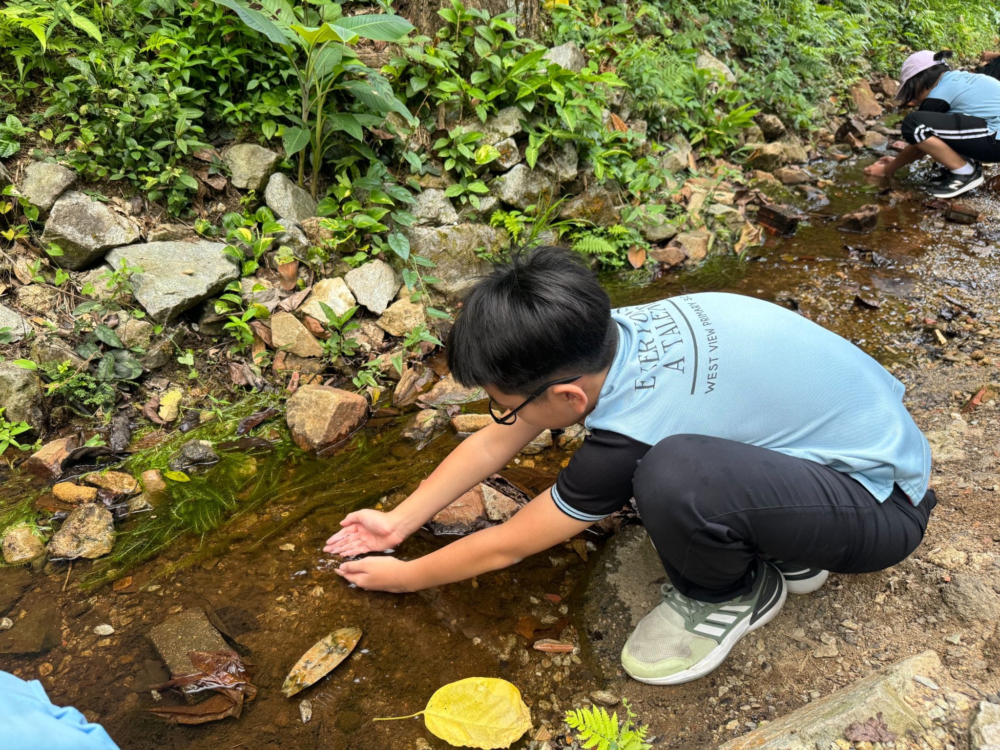 A boy in a light blue shirt crouches beside a rocky stream, scooping water with his hands outdoors.