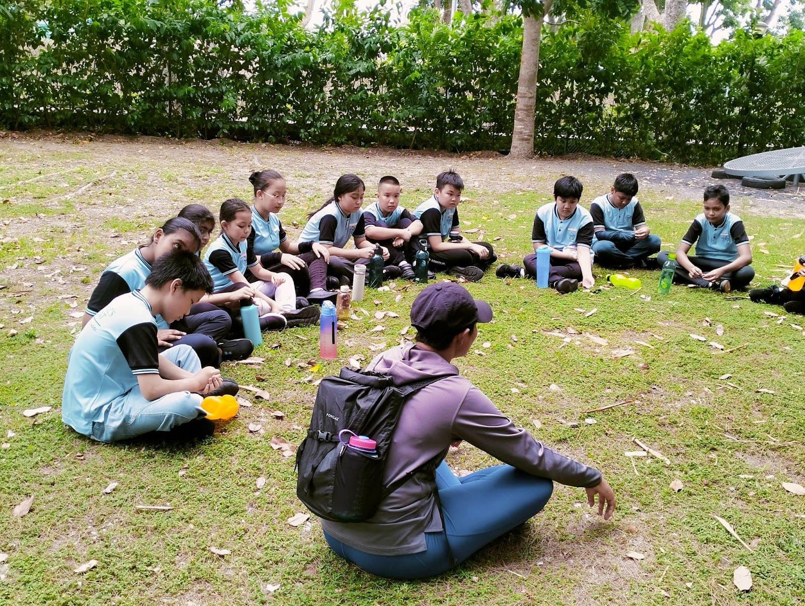 A adult with a backpack sits on grass facing a group of children in blue uniforms seated in a semicircle outdoors.