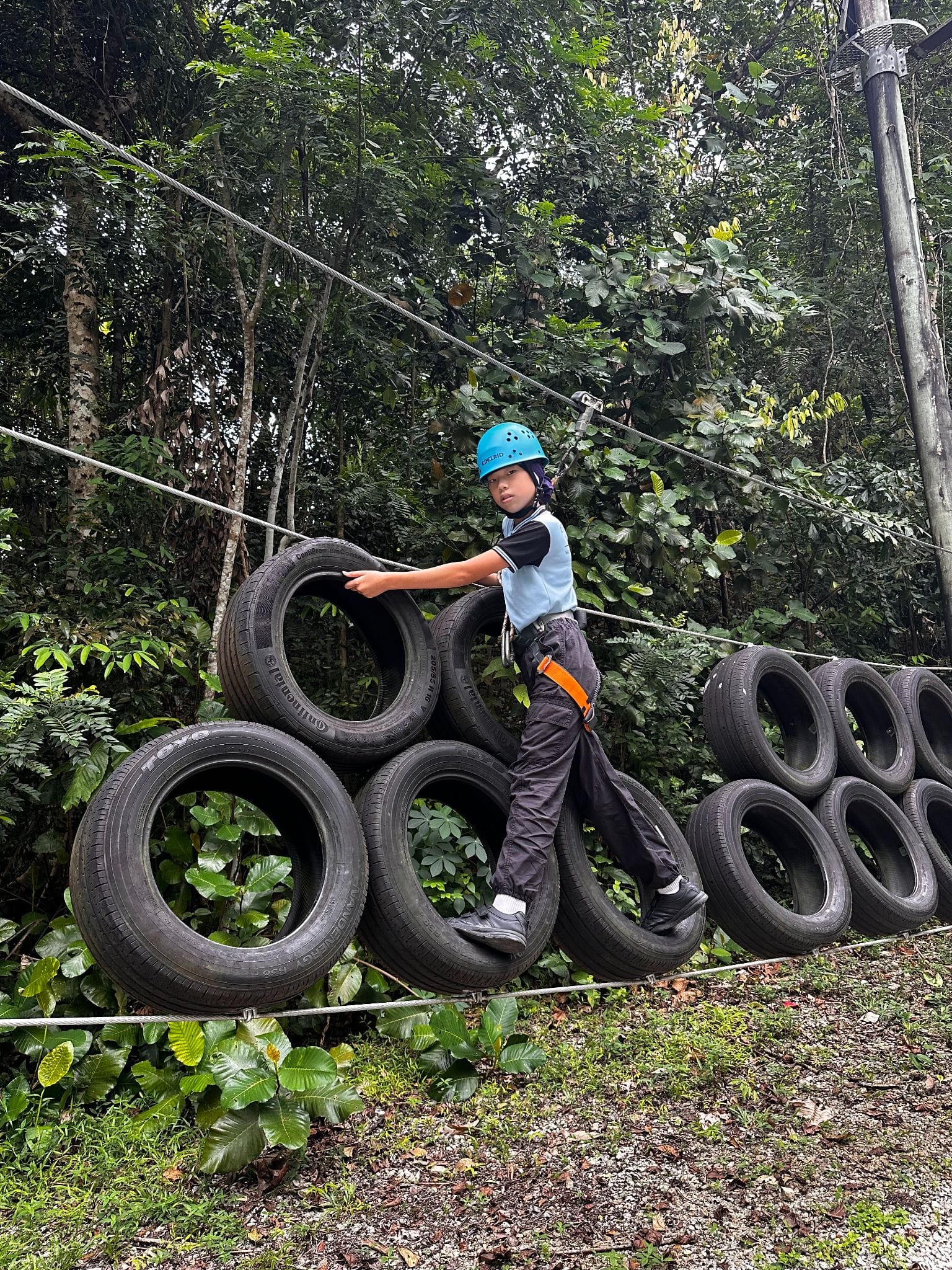 A child wearing a blue helmet and harness climbs over stacked tires on an outdoor obstacle course in a forest.