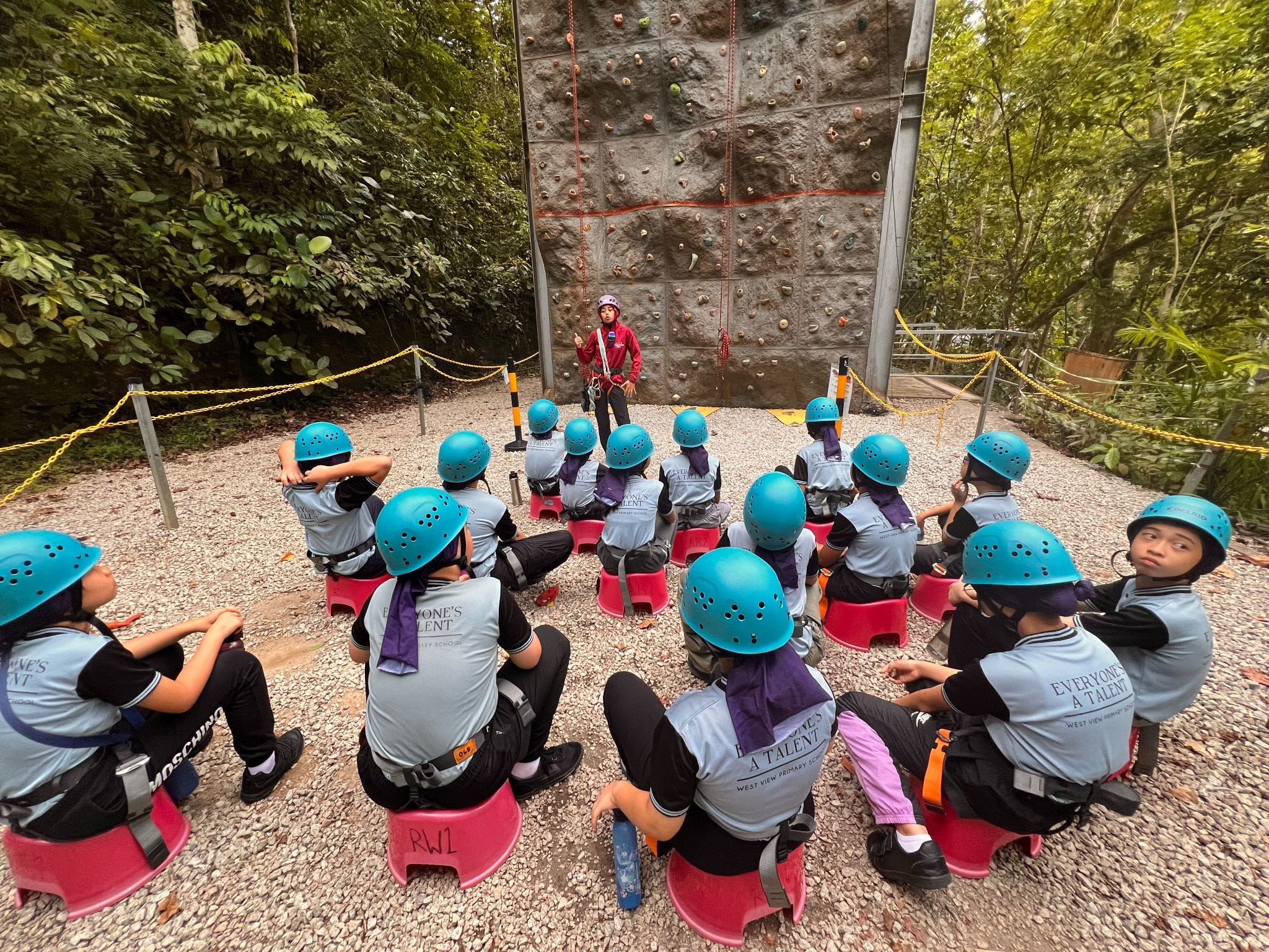 Students in blue helmets sit on red stools listening to an instructor in front of an outdoor rock climbing wall.
