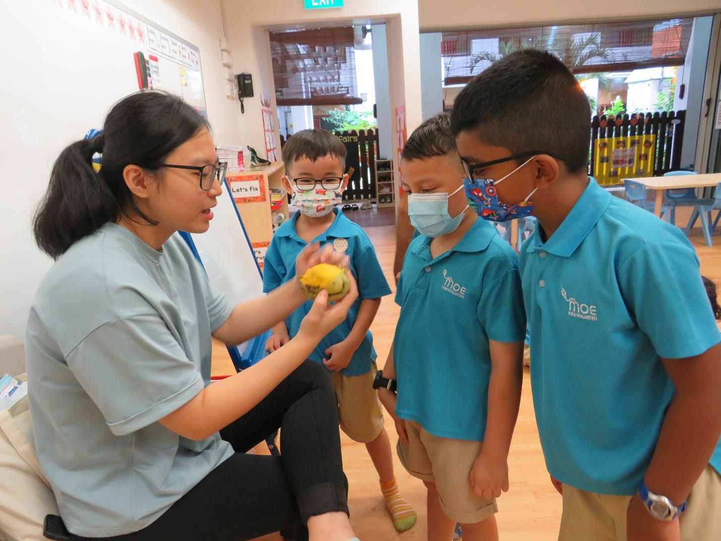A teacher holds a yellow fruit showing it to three young masked children in blue MOE Kindergarten uniforms indoors.