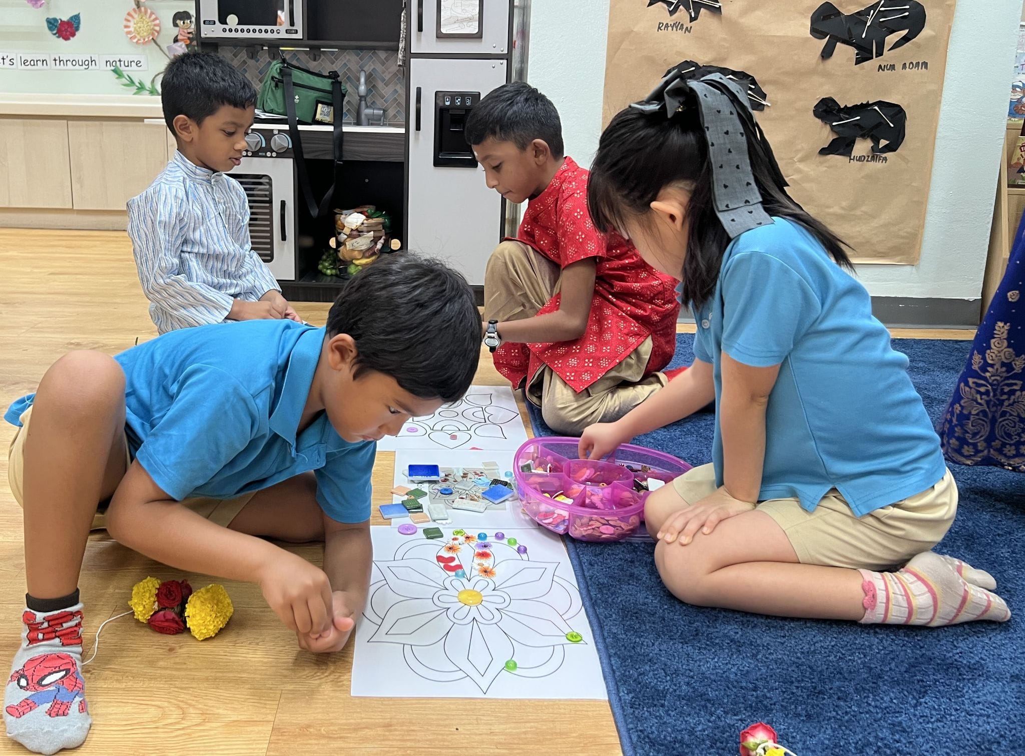 Four children sit on a classroom floor decorating a large floral mandala drawing with colorful beads and tiles.