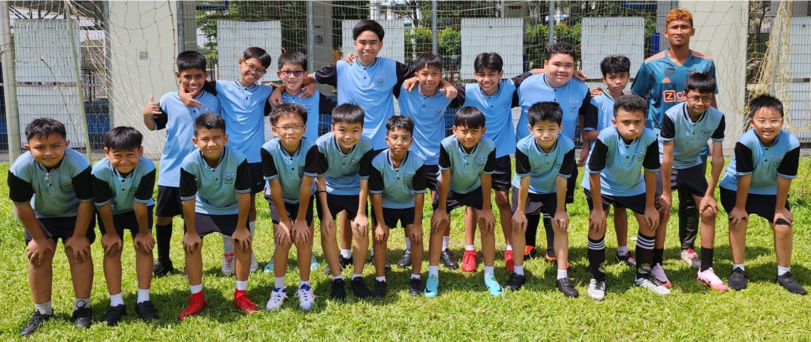 A youth soccer team of about 20 boys in light blue uniforms posing in two rows in front of a goal net.