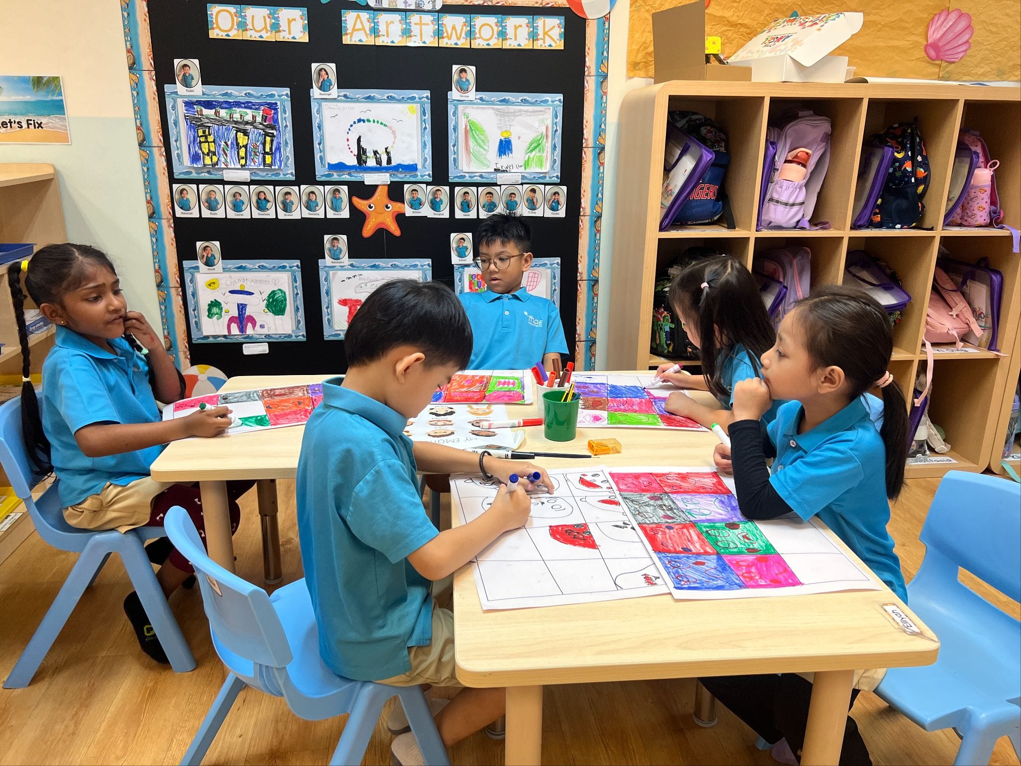 Five young students in blue uniforms draw and color at tables in a classroom with an "Our Artwork" bulletin board behind them.