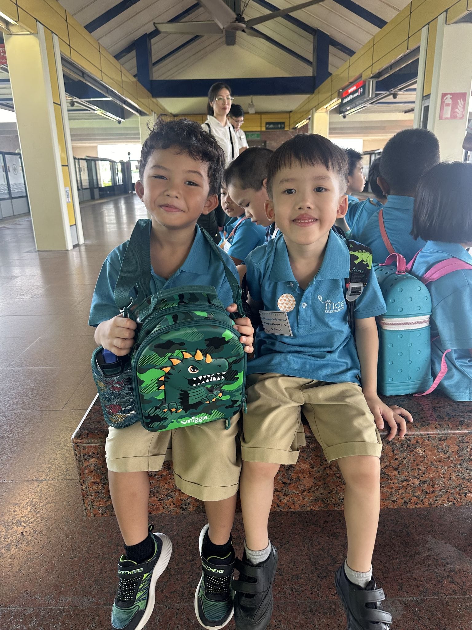 Two young boys in blue school uniforms sit together at a transit station, one holding a green dinosaur backpack.