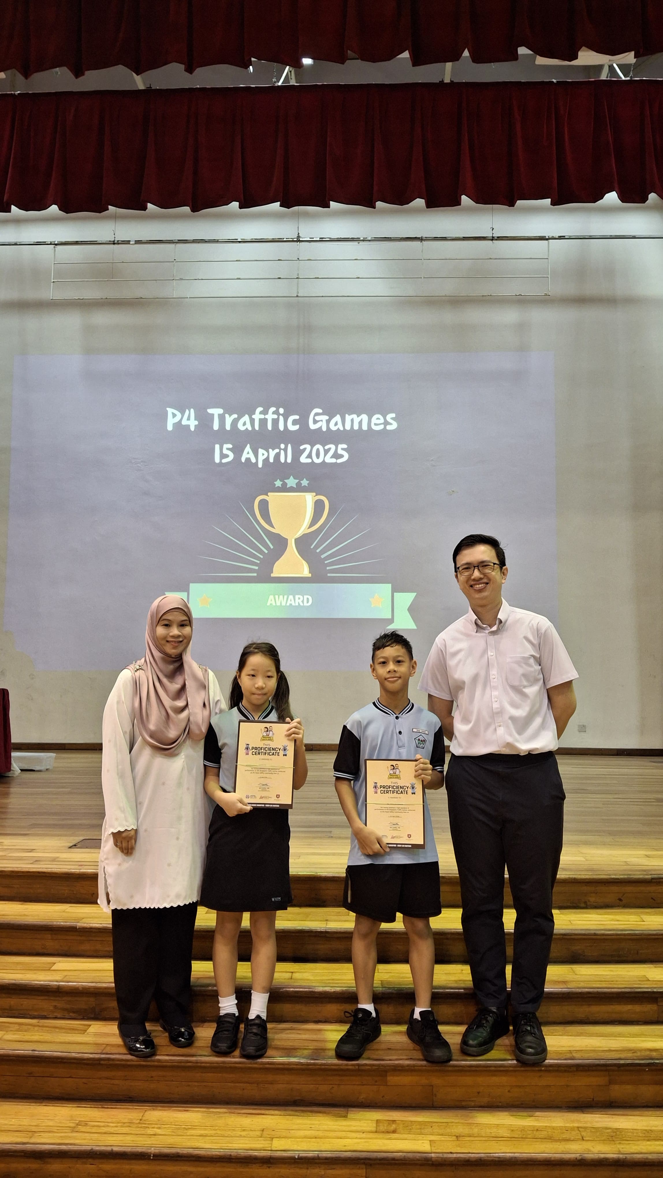 Two adults and two children holding proficiency certificates on a stage at P4 Traffic Games, 15 April 2025.