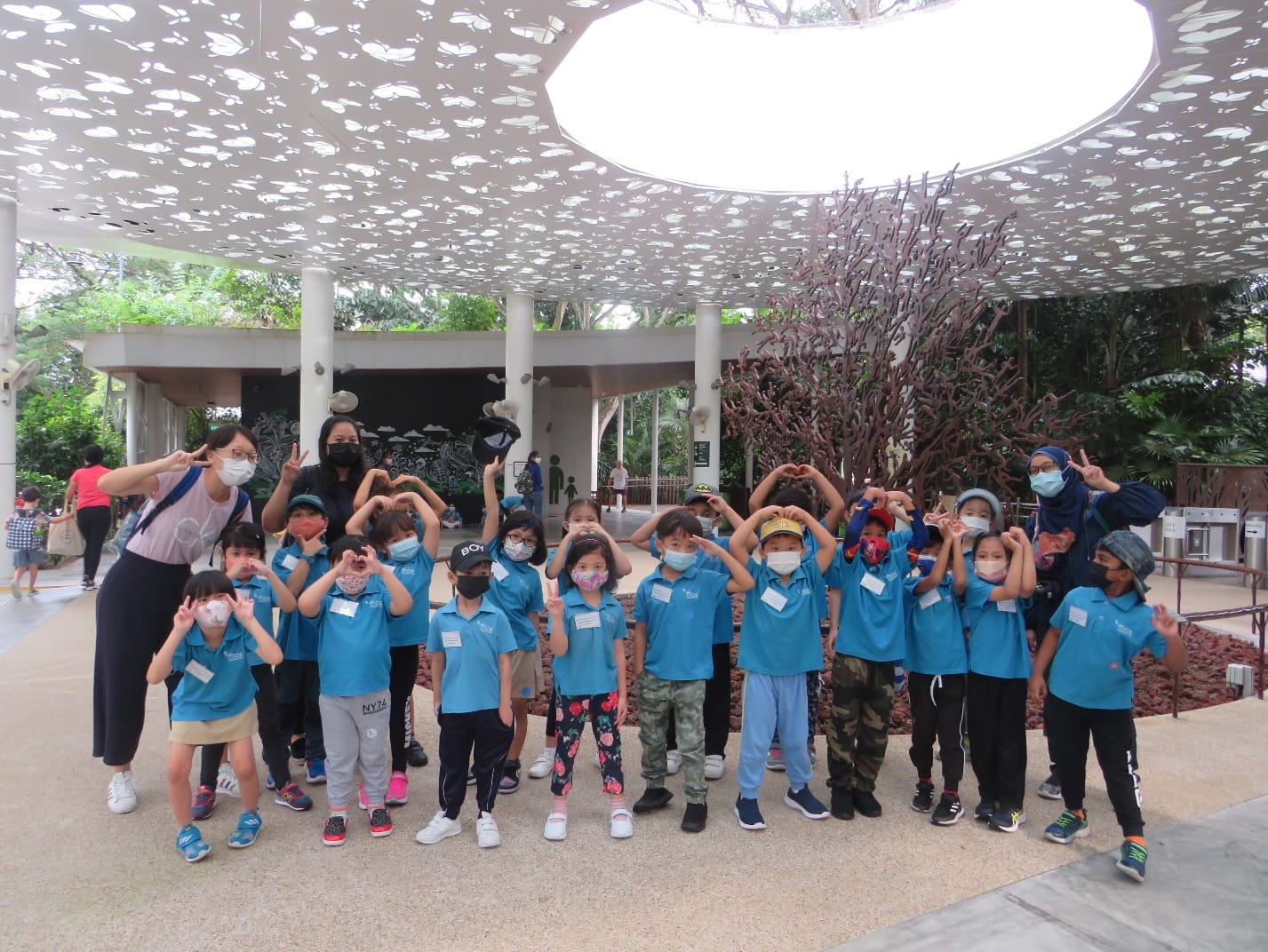 A group of young children in blue shirts and masks pose with heart hand gestures under a decorative canopy outdoors.