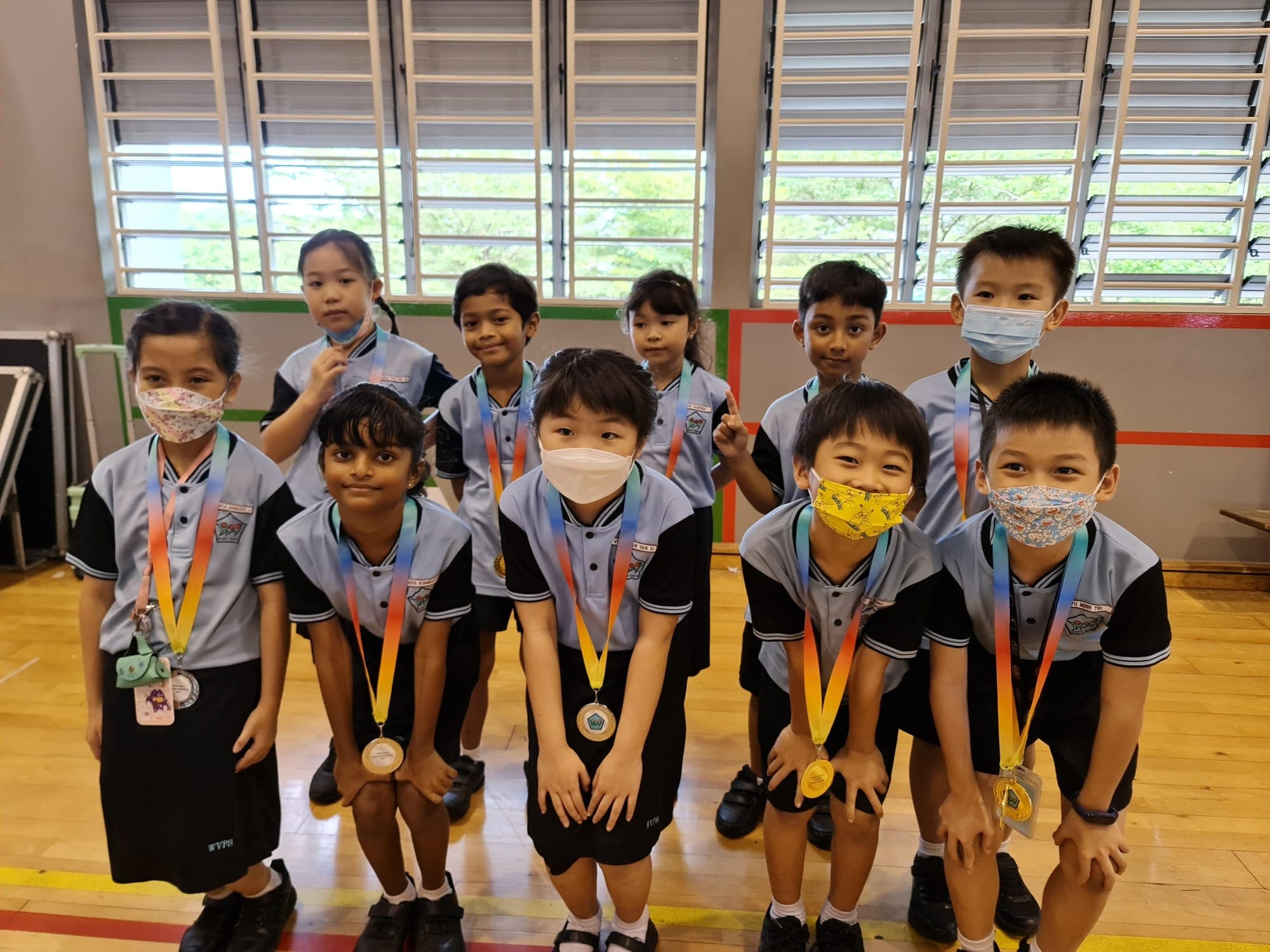 Ten young students in school uniforms and face masks pose indoors wearing colorful medal ribbons around their necks.