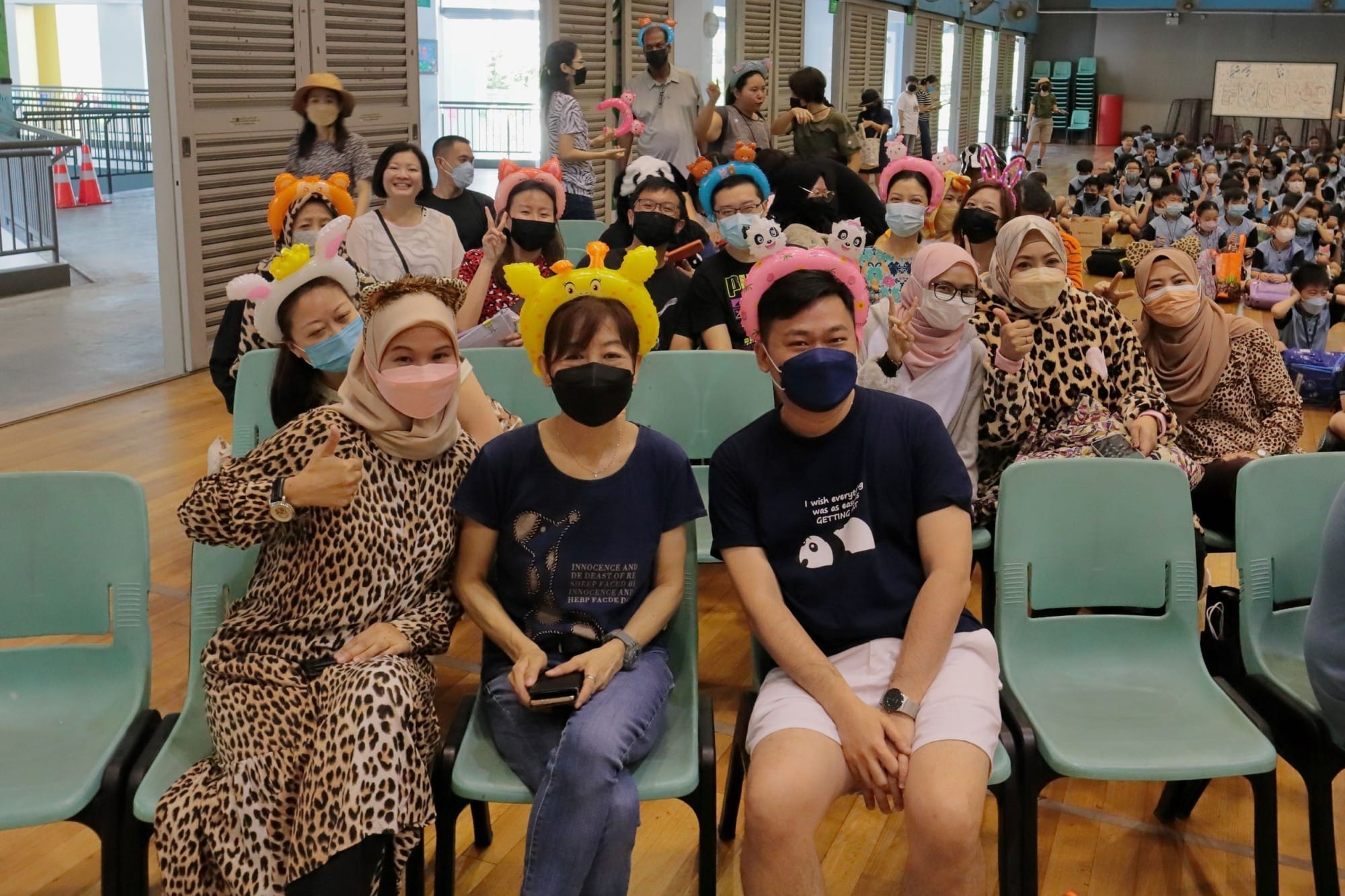 A large group of masked adults wearing novelty animal headbands sit on chairs in a school hall, smiling at the camera.