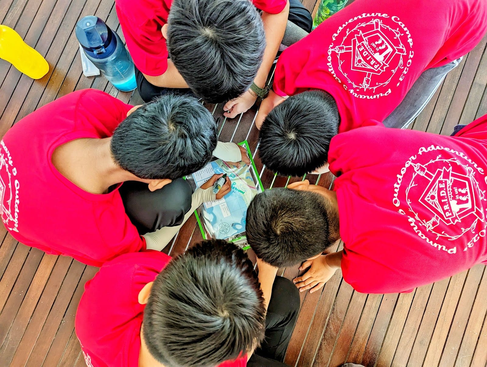 Several children in red Outdoor Adventure Camp shirts huddle over a map on a wooden deck.