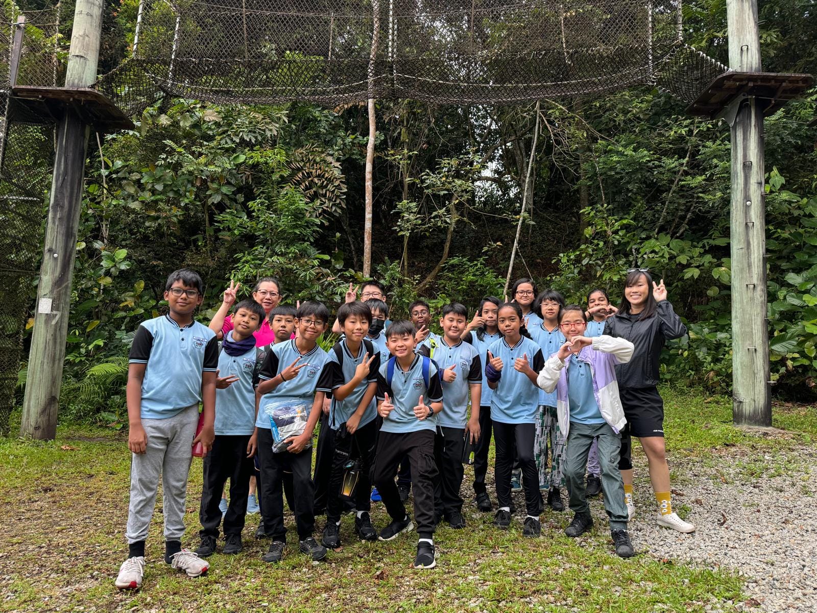 A group of students in light blue uniforms poses outdoors near a large net structure surrounded by trees.