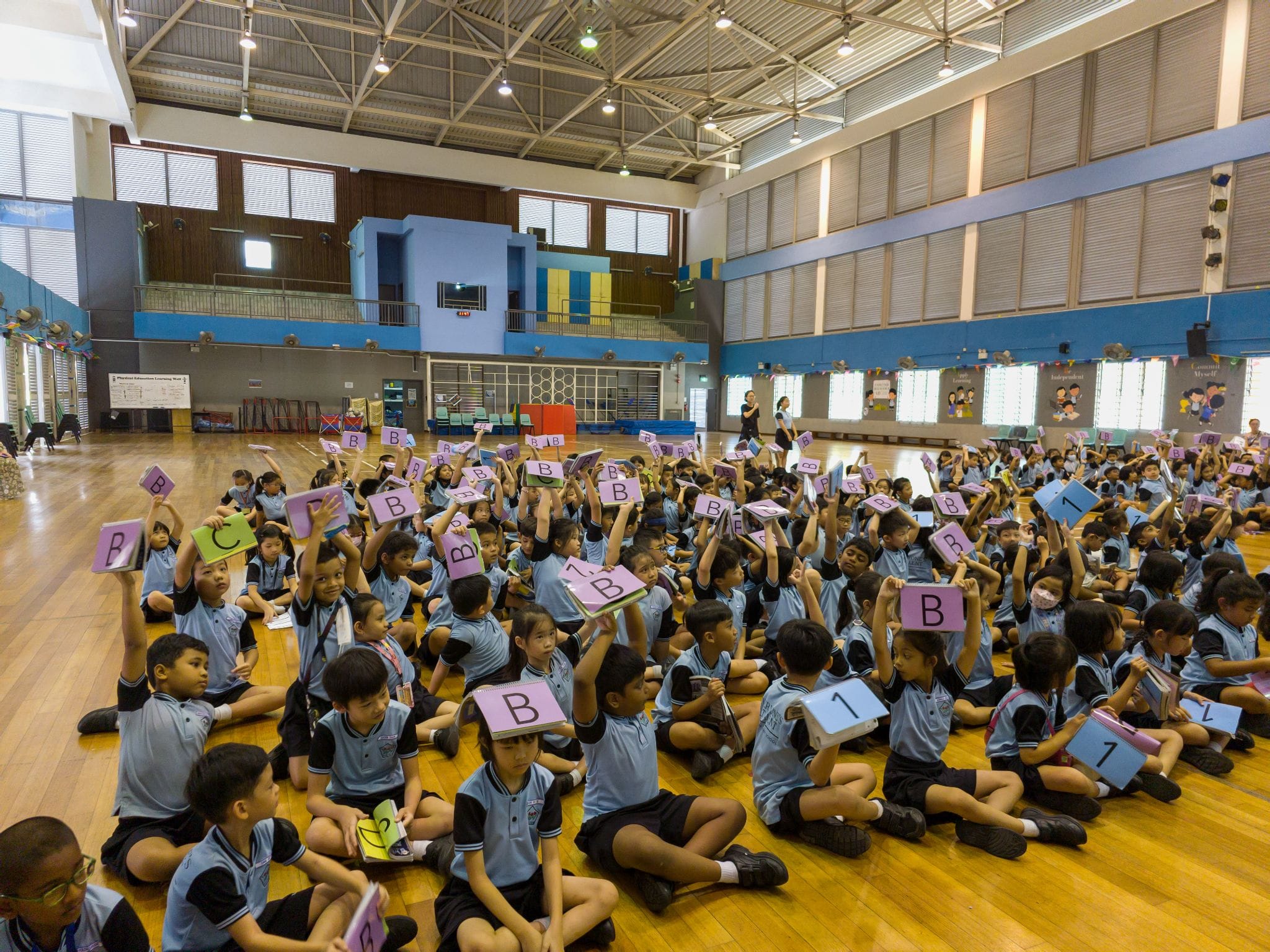 Large group of young students in blue uniforms sitting on a gym floor, holding up colorful cards with letters and numbers.