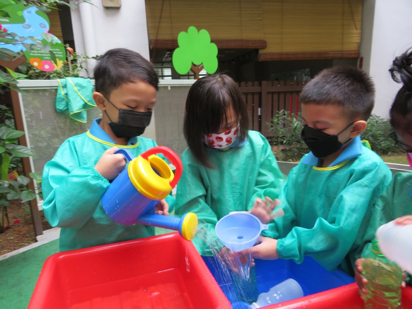 Three masked children in teal aprons play with a toy watering can and cups at a red water table outdoors.