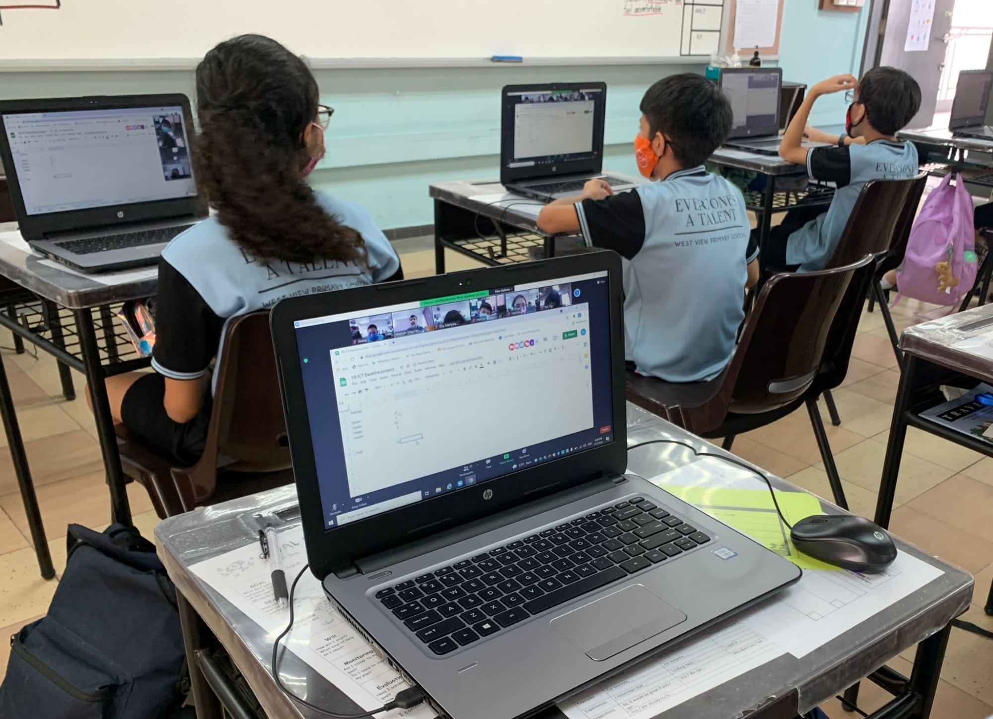 Three masked students in school uniforms sit at desks using HP laptops showing a video call in a classroom.