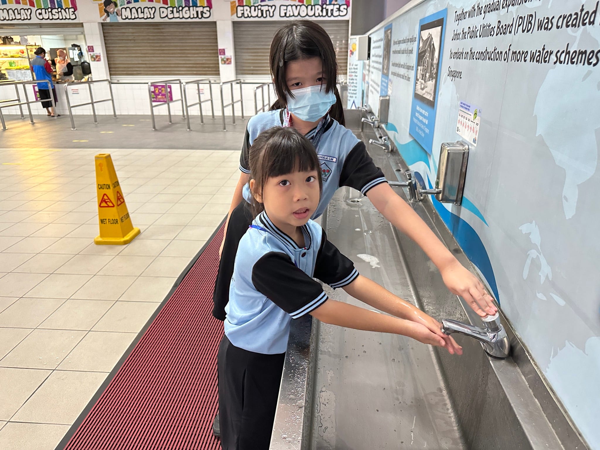 Two young girls in school uniforms wash hands at a long metal sink inside a food court.
