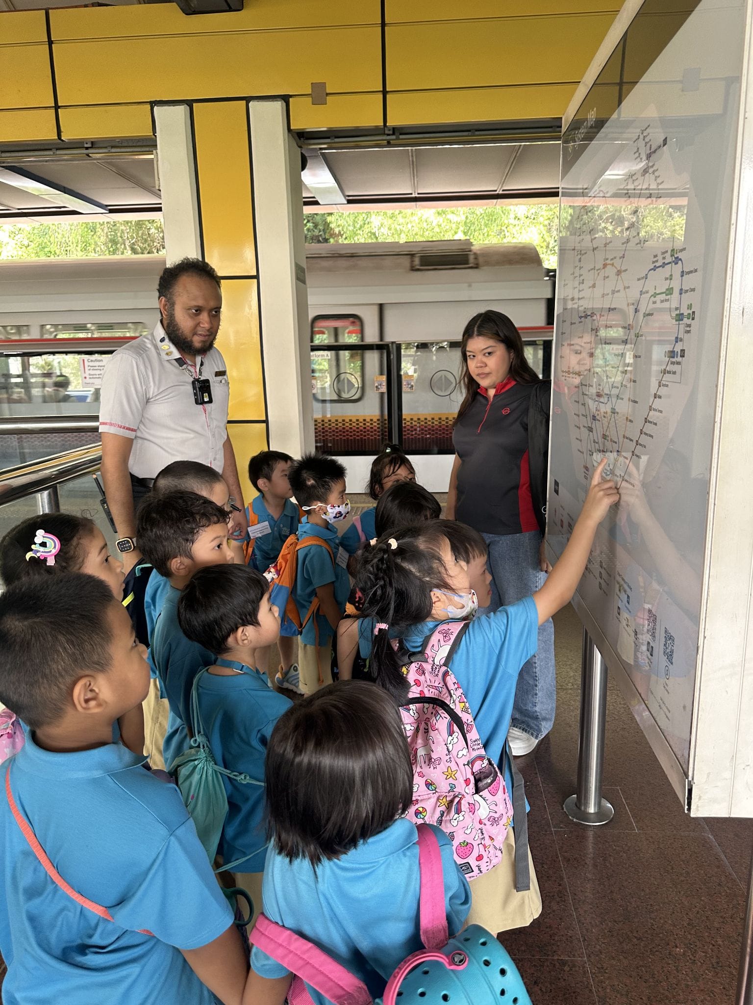 A woman points at a transit map while a group of young children in blue uniforms gathers around at a train station.
