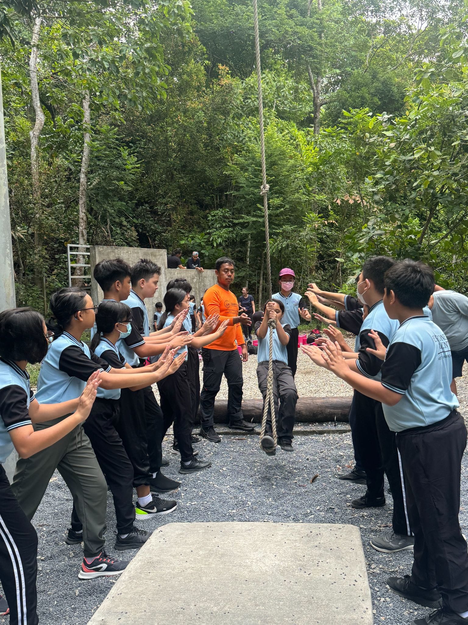 Students in light blue uniforms clap as a peer grabs a hanging rope during an outdoor team activity in a forest.