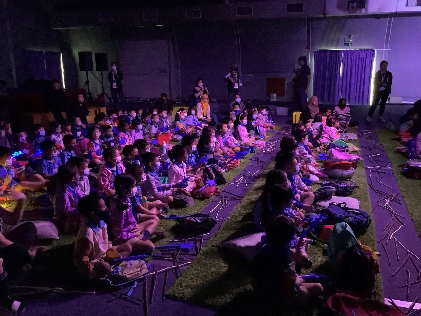 A large group of children sit on mats on a floor in a dimly lit hall, facing a stage with purple lighting.