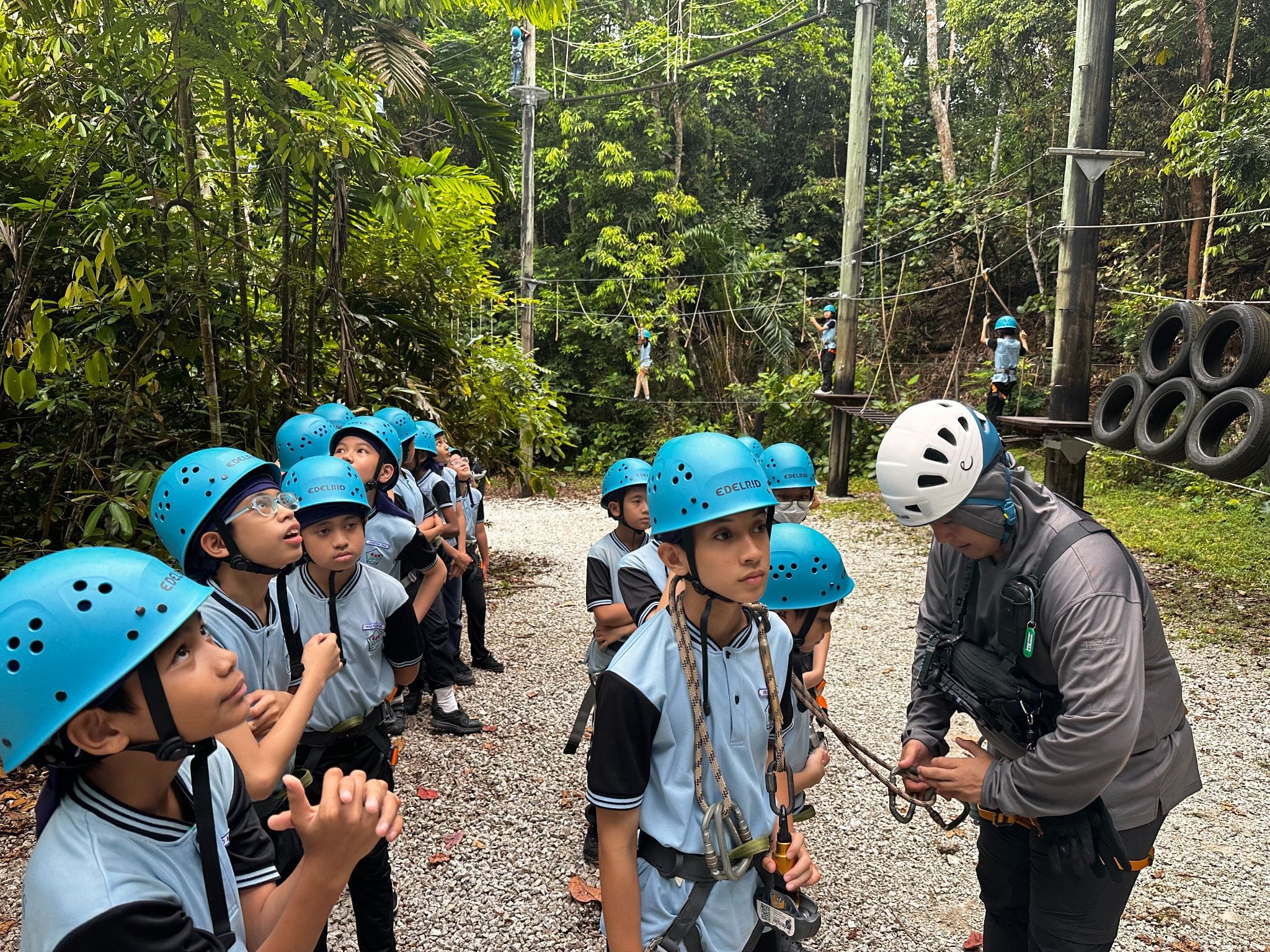 Students in blue helmets and harnesses listen to an instructor at an outdoor ropes course surrounded by forest.