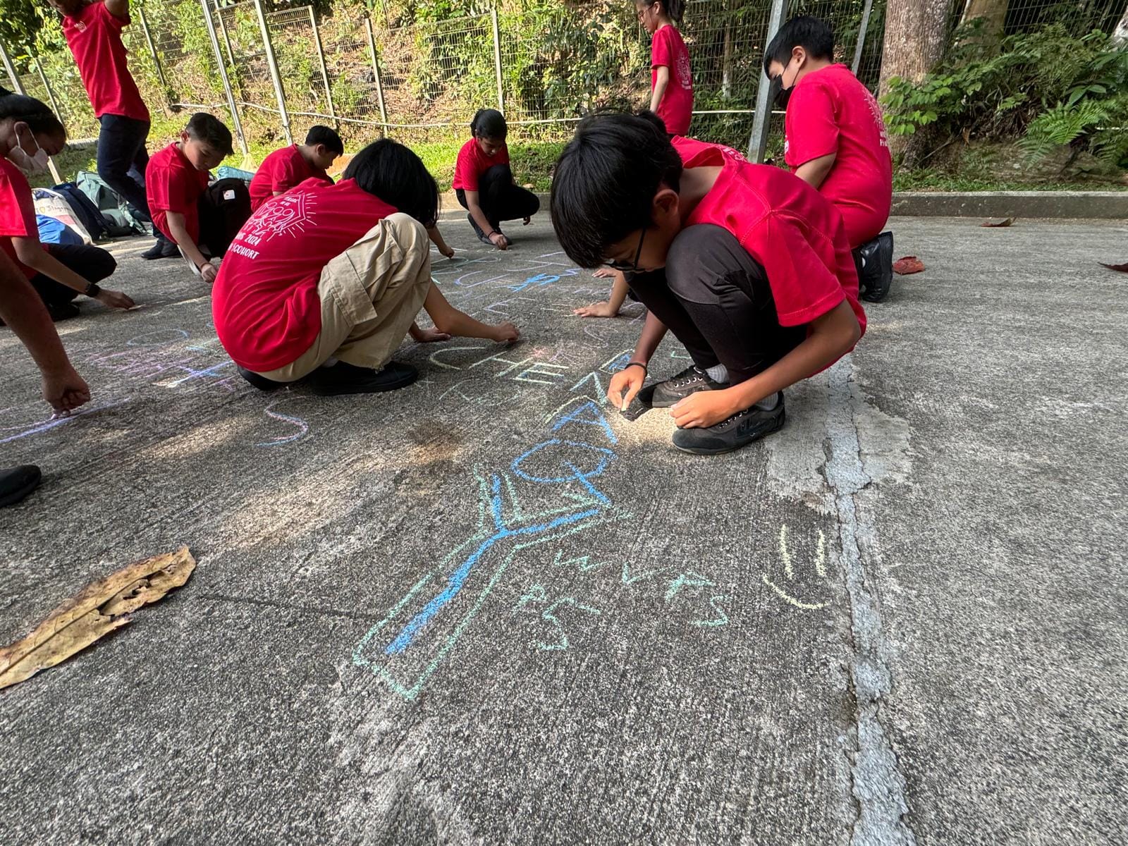 Children in red shirts crouch on pavement drawing colorful chalk letters and designs outdoors near a fence.