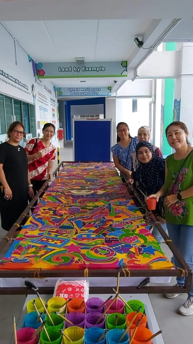 Five women stand around a large colorful batik fabric on a frame, with paint cups and brushes in the foreground.