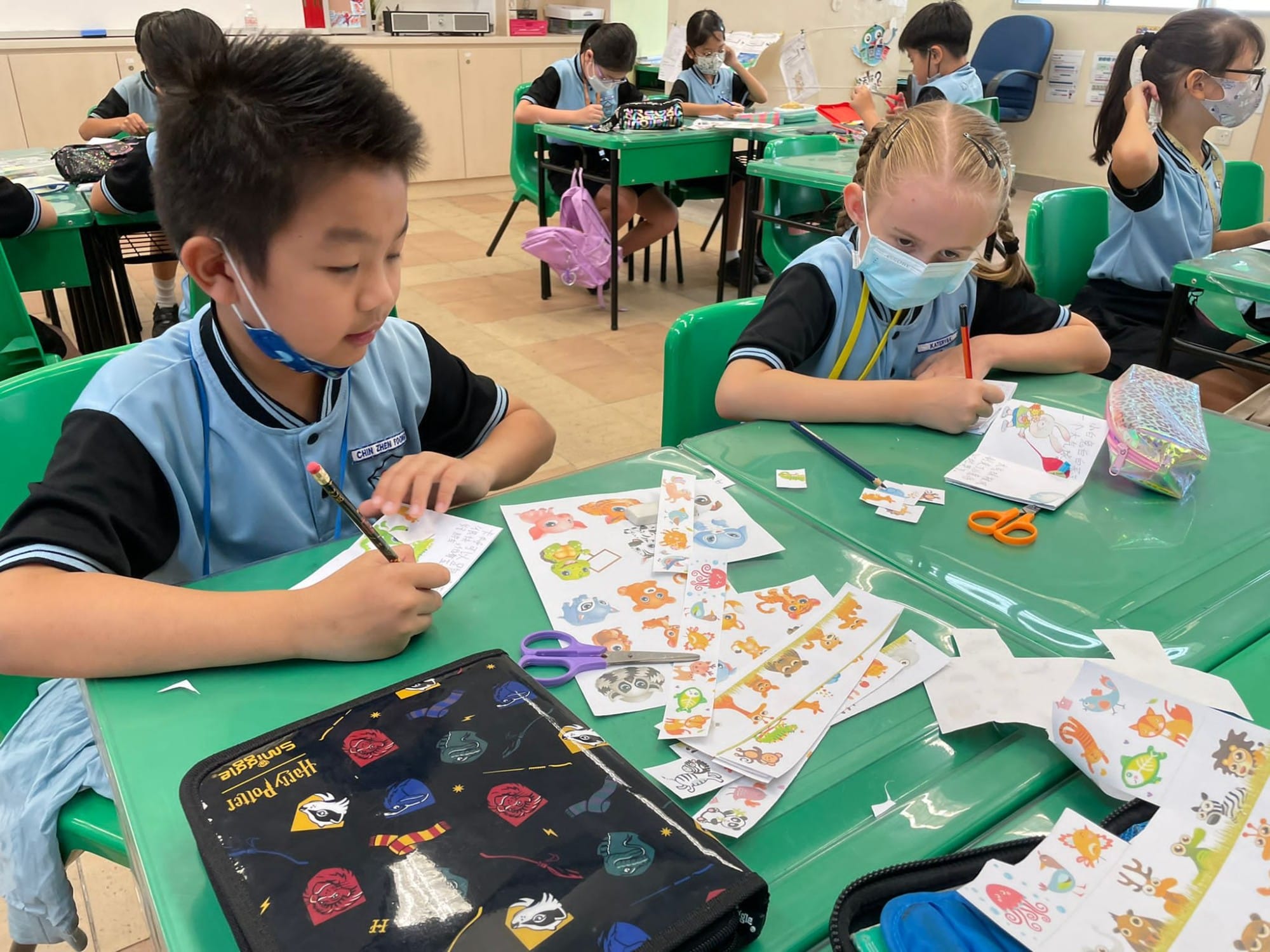 Two masked children in blue school uniforms sit at a green desk cutting colorful animal stickers in a classroom.
