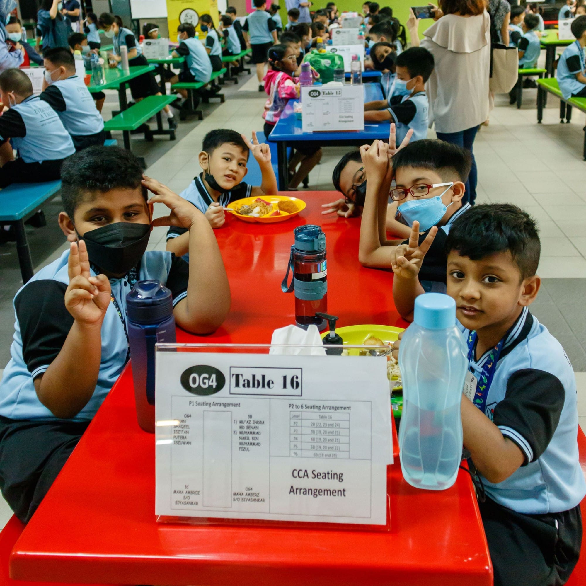 Young students in blue uniforms sit at a red cafeteria table labeled OG4 Table 16, making peace signs.