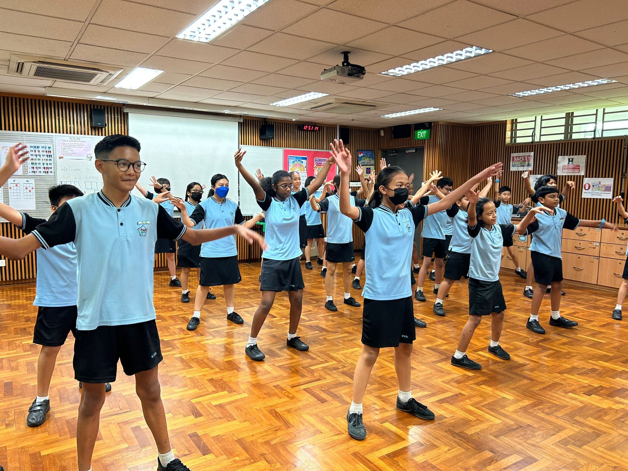 Students in light blue and black uniforms practice a dance routine with raised arms in a wood-floored school room.