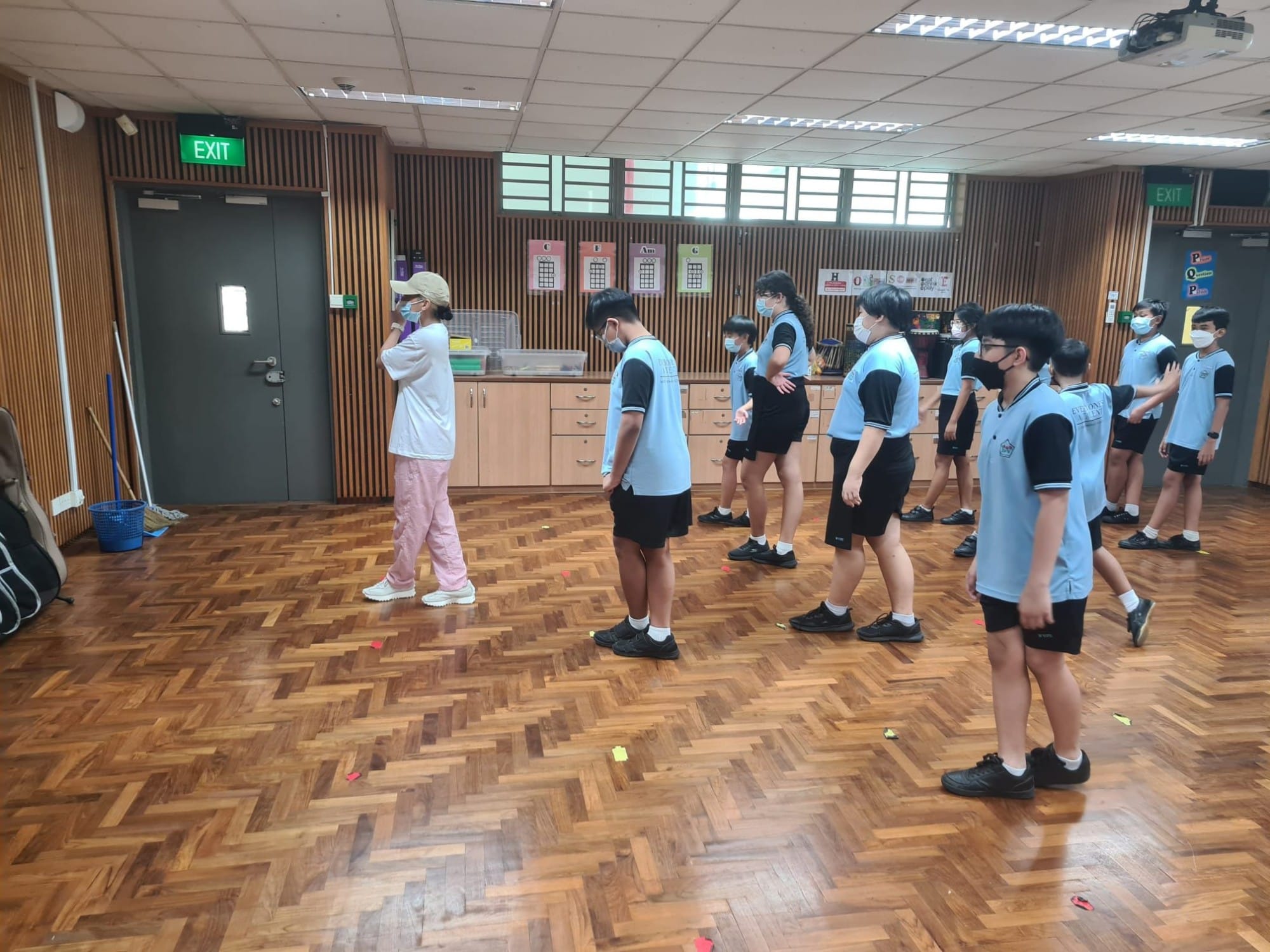 Students in light blue uniforms and masks stand in rows on a wooden floor in an indoor hall with exit signs.