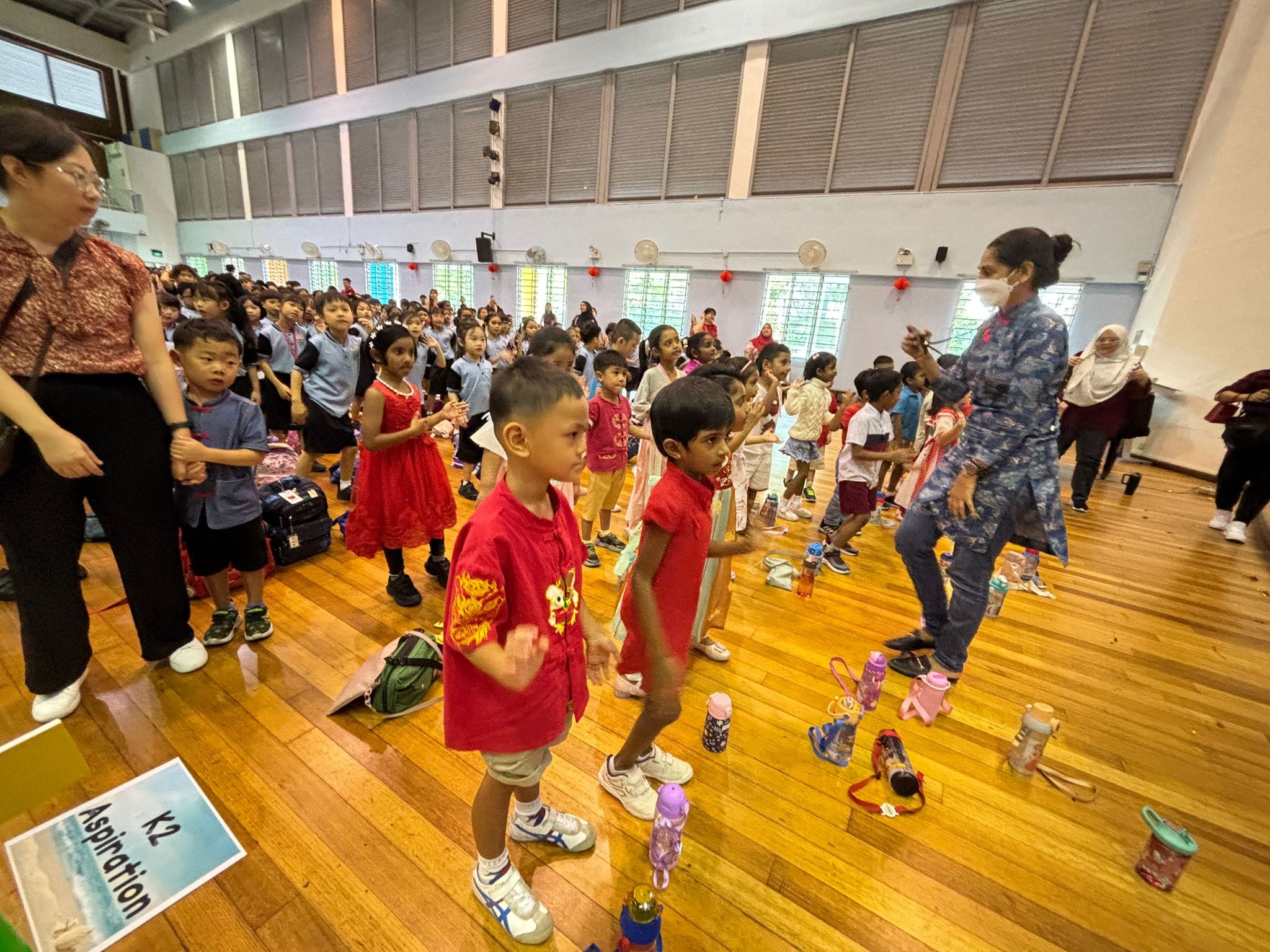 Young children in red and blue uniforms stand in lines on a wooden hall floor, guided by adult teachers.
