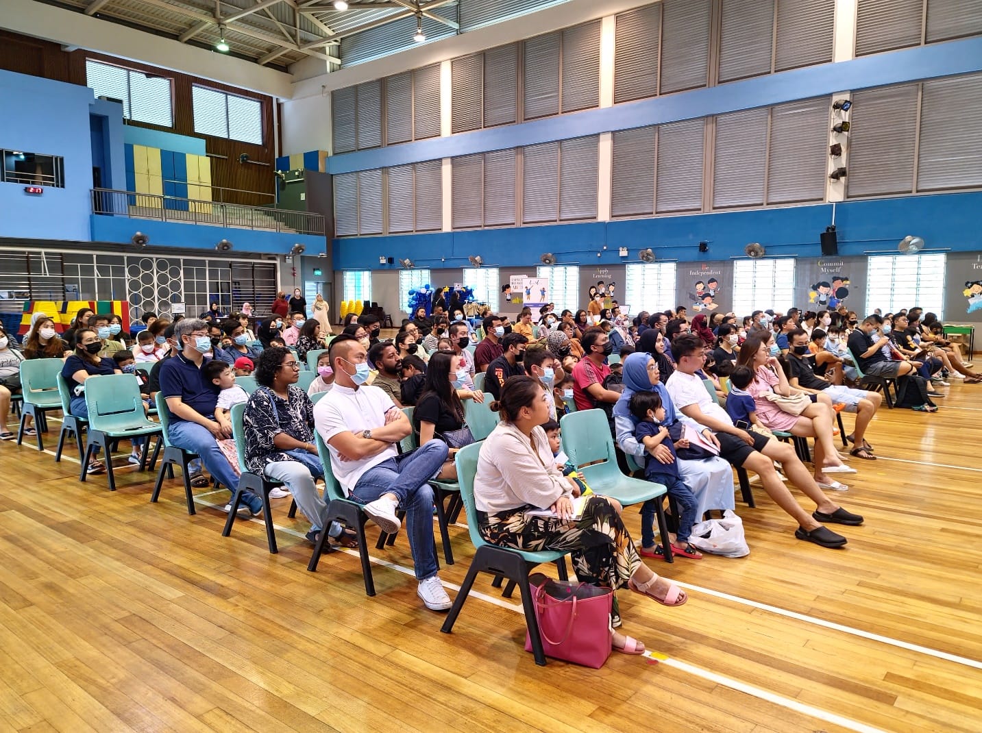 A large crowd of adults and children seated on teal chairs in a school hall, facing a stage at the front.