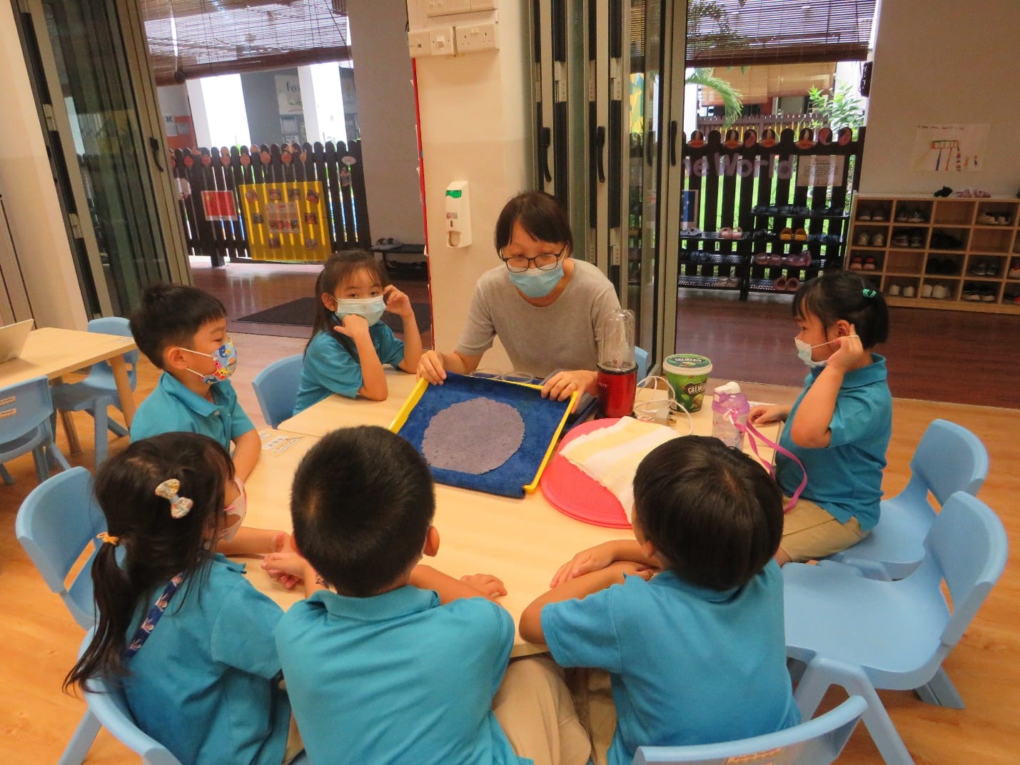 A masked teacher and six young children in teal uniforms sit around a table with a sand tray in a classroom.