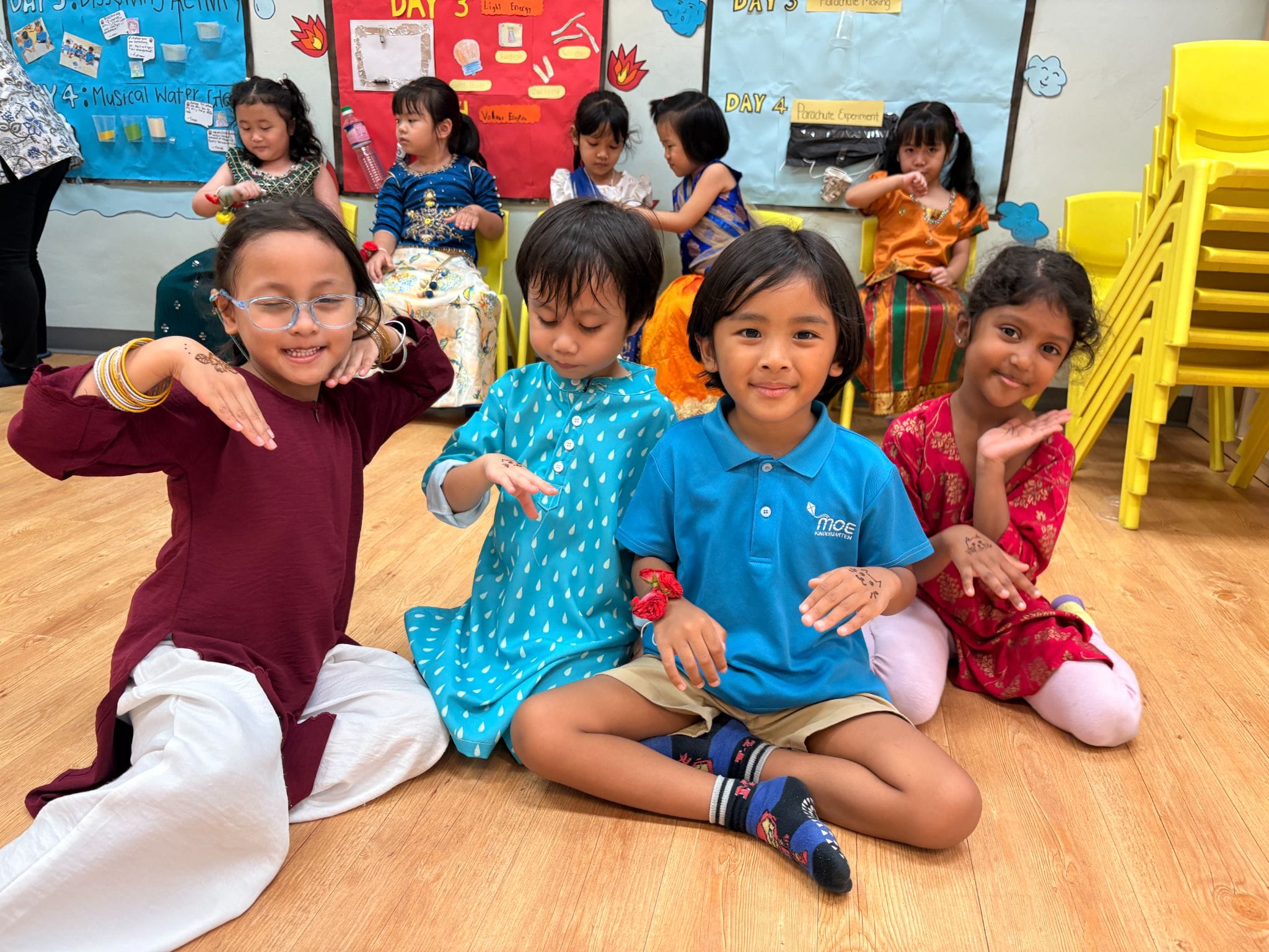 Four young children in traditional and school attire sit on a classroom floor, smiling and showing henna on their hands.