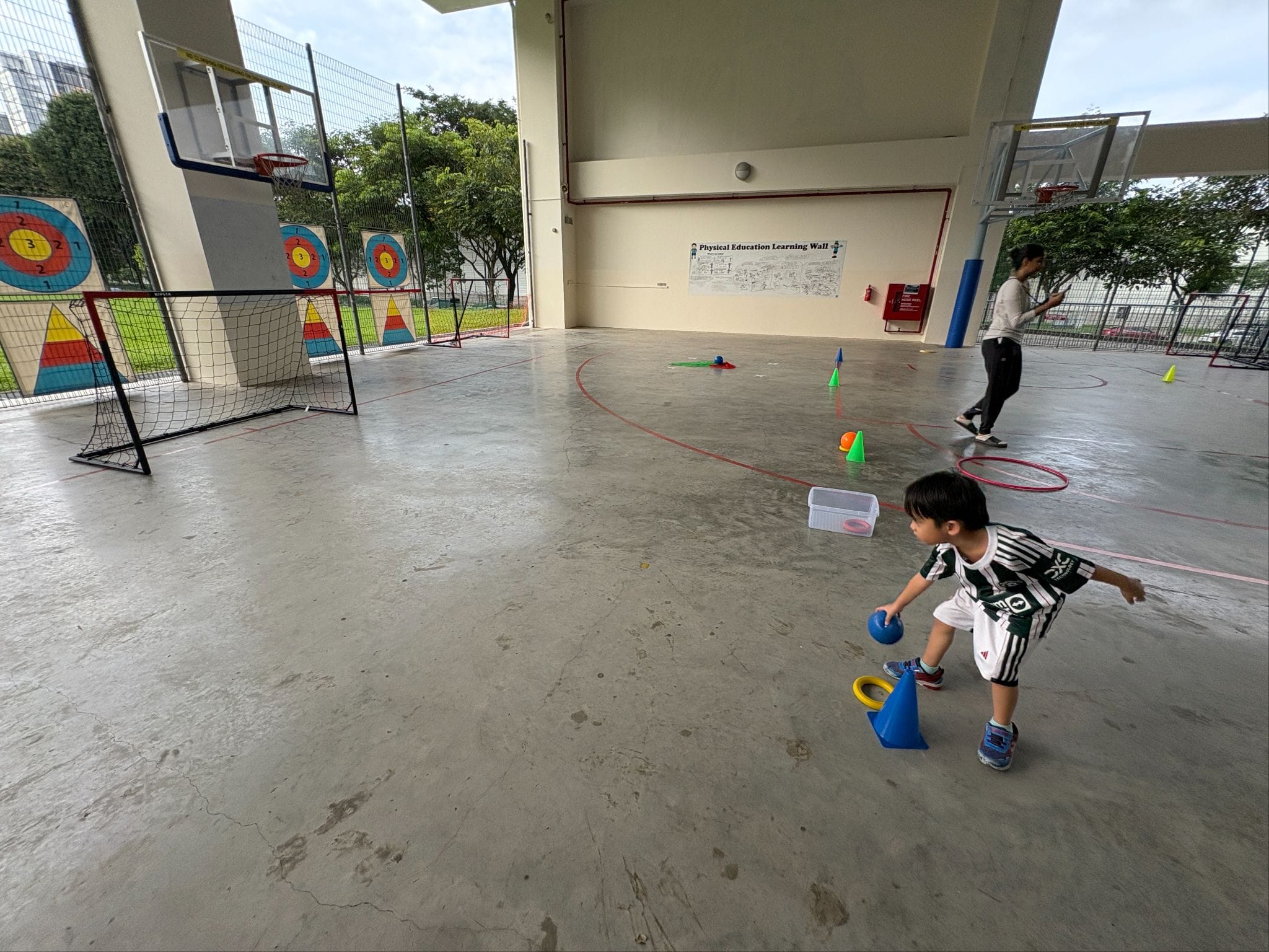 A young child picks up a blue ball near cones on a covered school sports court with archery targets and a soccer goal.
