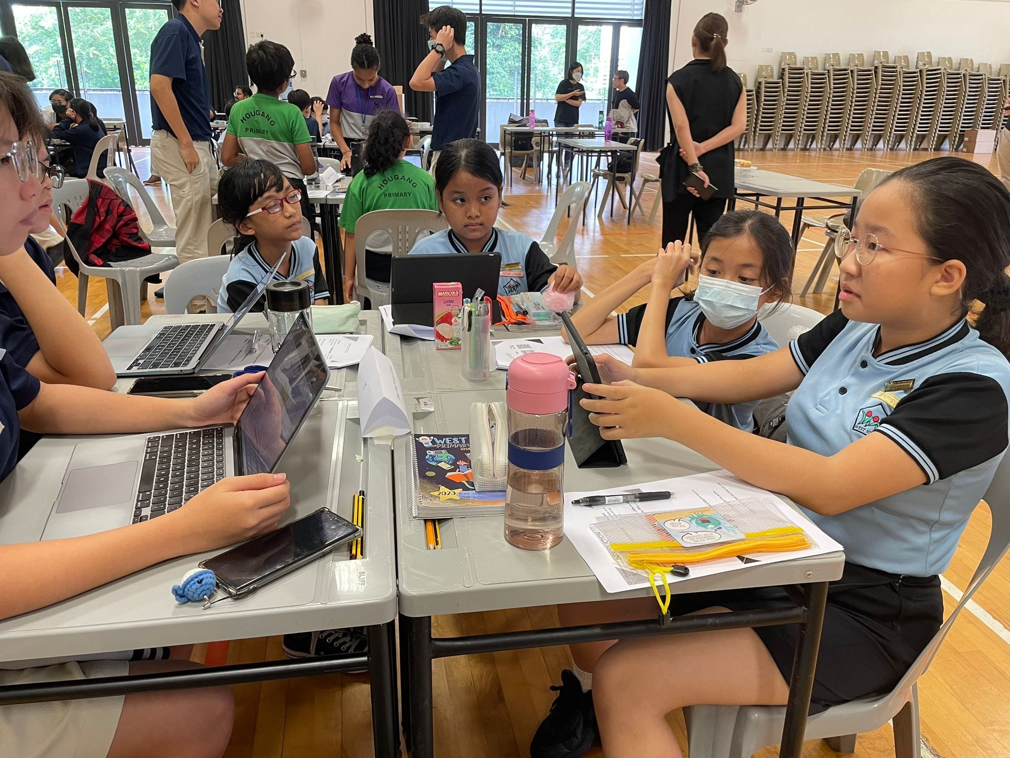 Primary school students in uniforms collaborate around a table with laptops and tablets in a school hall.
