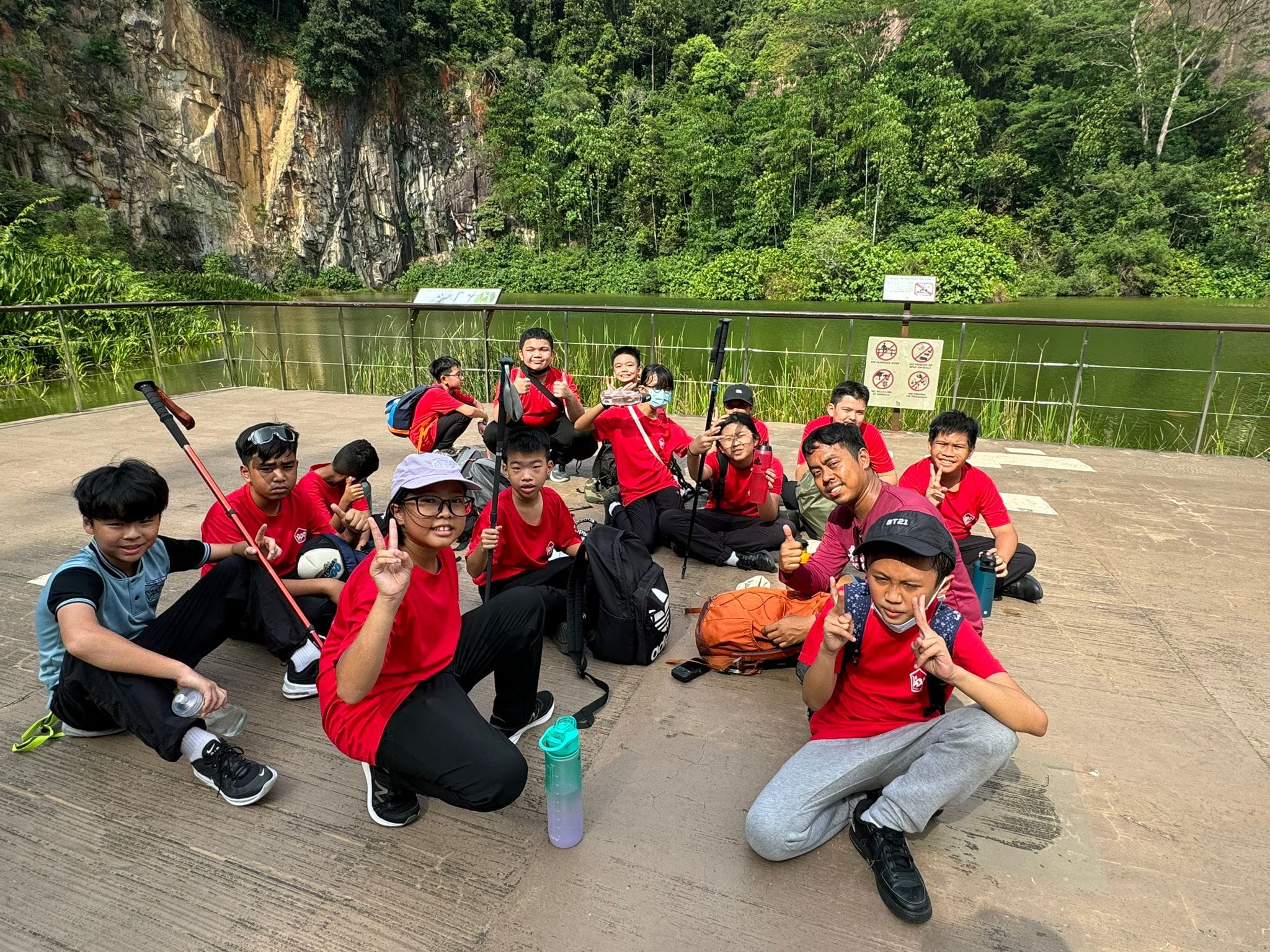 A group of kids in red shirts pose and smile on a platform near a green lake with rocky cliffs and trees behind them.