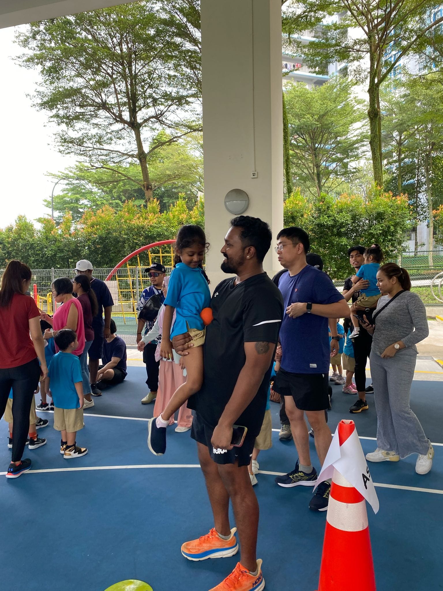 A man in black holds a young girl in blue on an outdoor court surrounded by families and trees.
