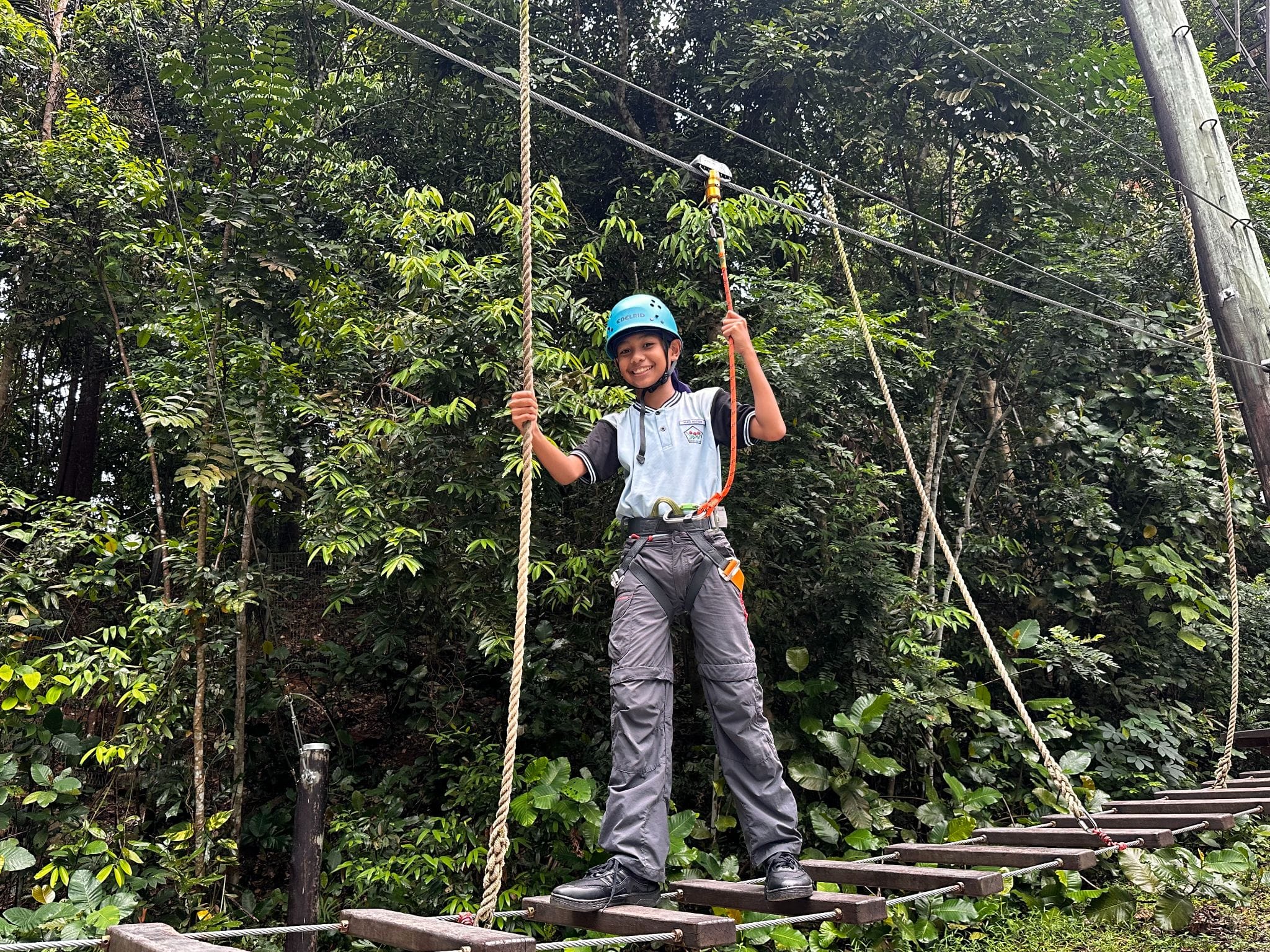 A smiling child wearing a blue helmet and harness stands on a rope bridge course surrounded by green trees.