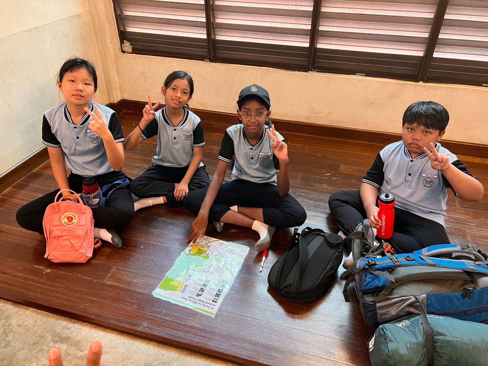 Four children in matching uniforms sit on a wooden floor with backpacks and a hiking trail map, making peace signs.