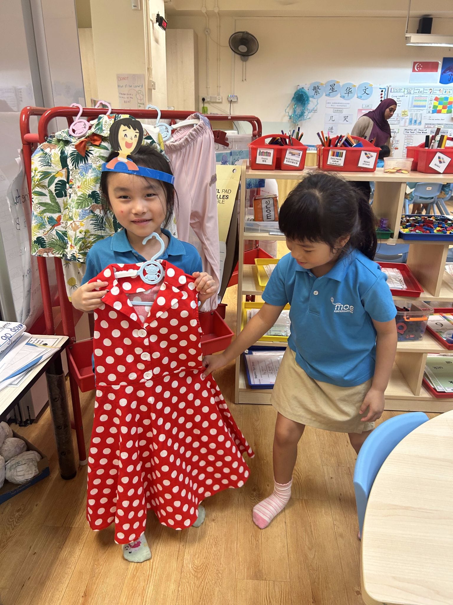 Two young girls in blue uniforms in a classroom, one holding a red polka-dot dress on a hanger.