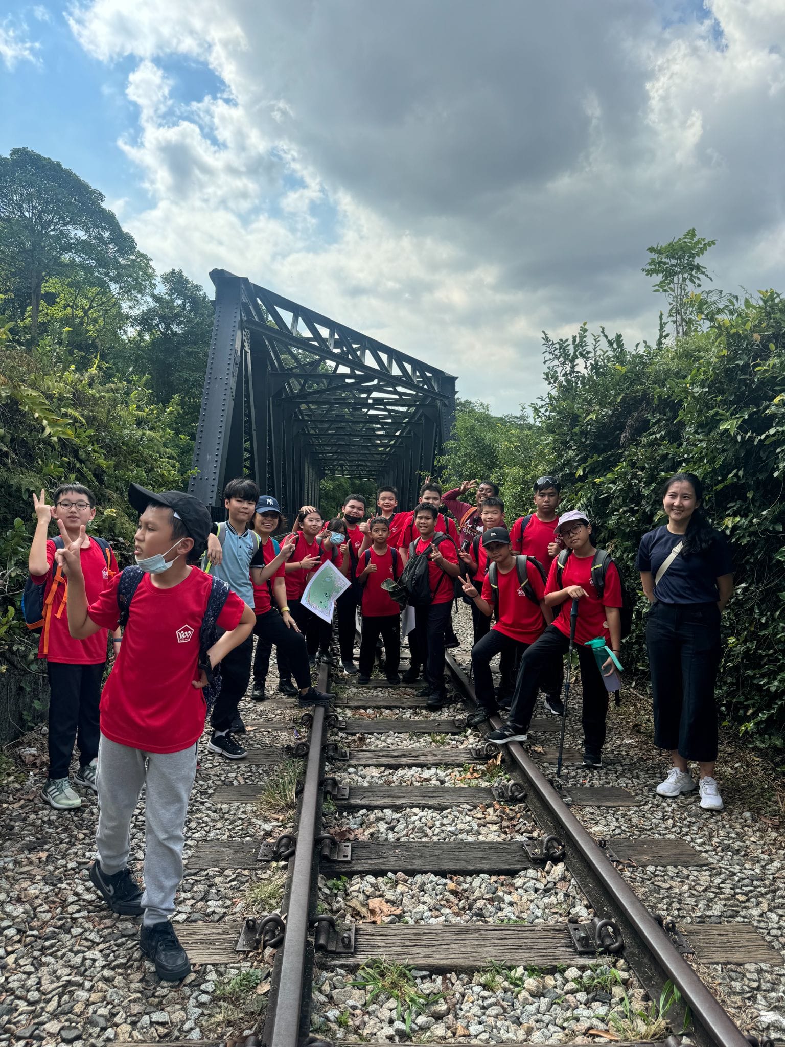 A group of children in red shirts pose on railway tracks in front of a steel truss bridge surrounded by trees.