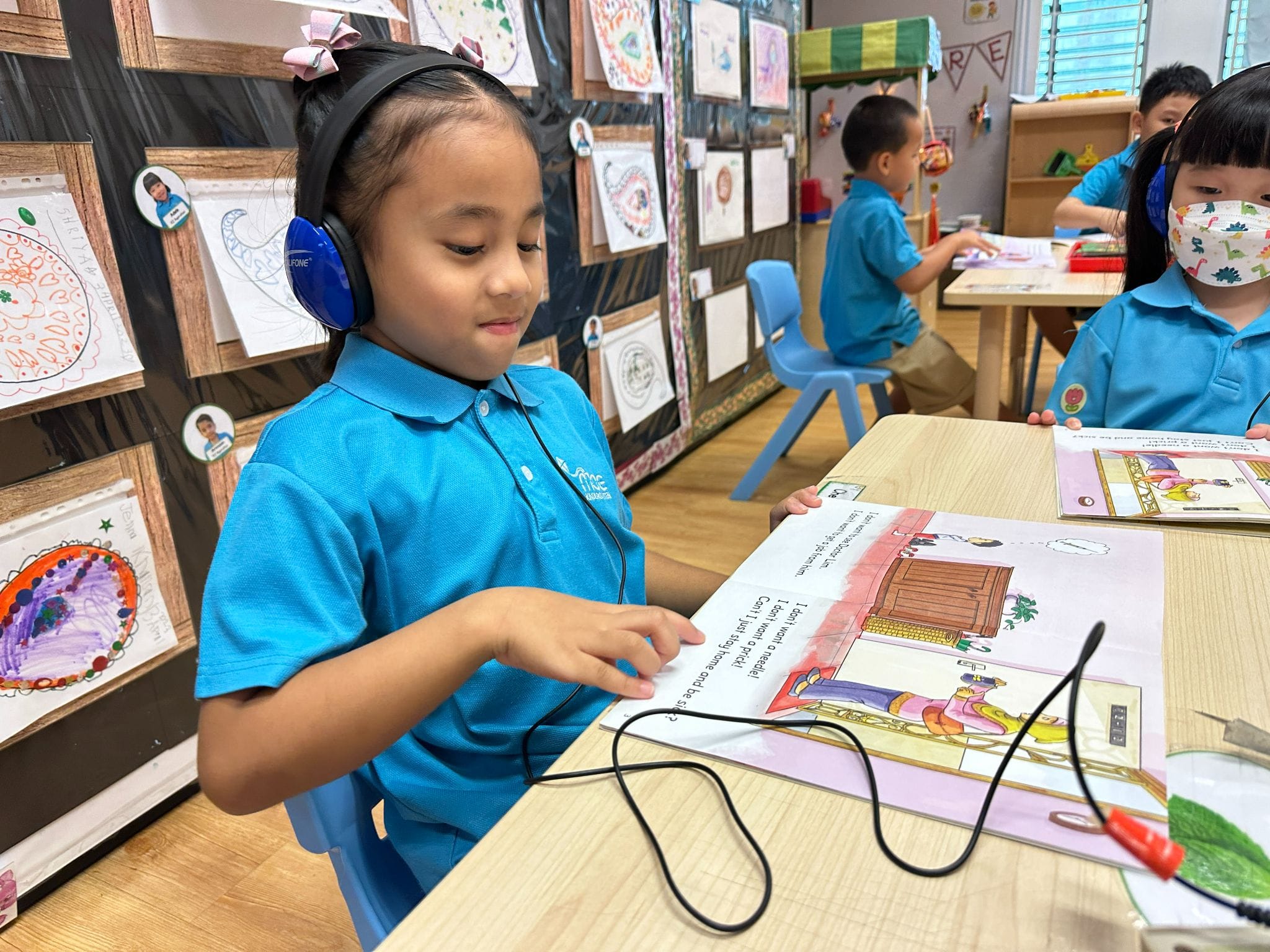 Young girl wearing blue headphones reads a picture book at a classroom desk, with other children and artwork visible behind her.