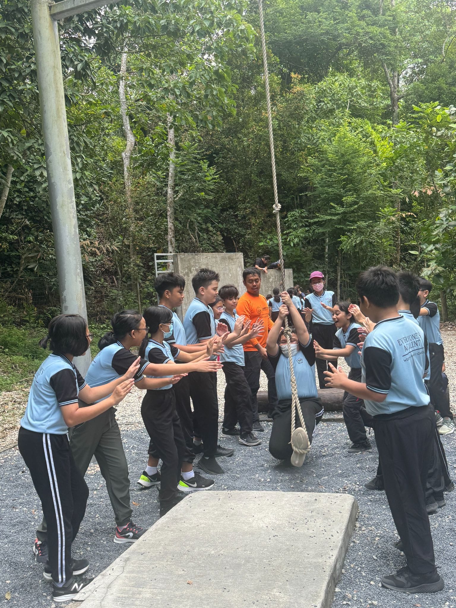Students in light blue uniforms gather around a hanging rope at an outdoor obstacle course surrounded by trees.