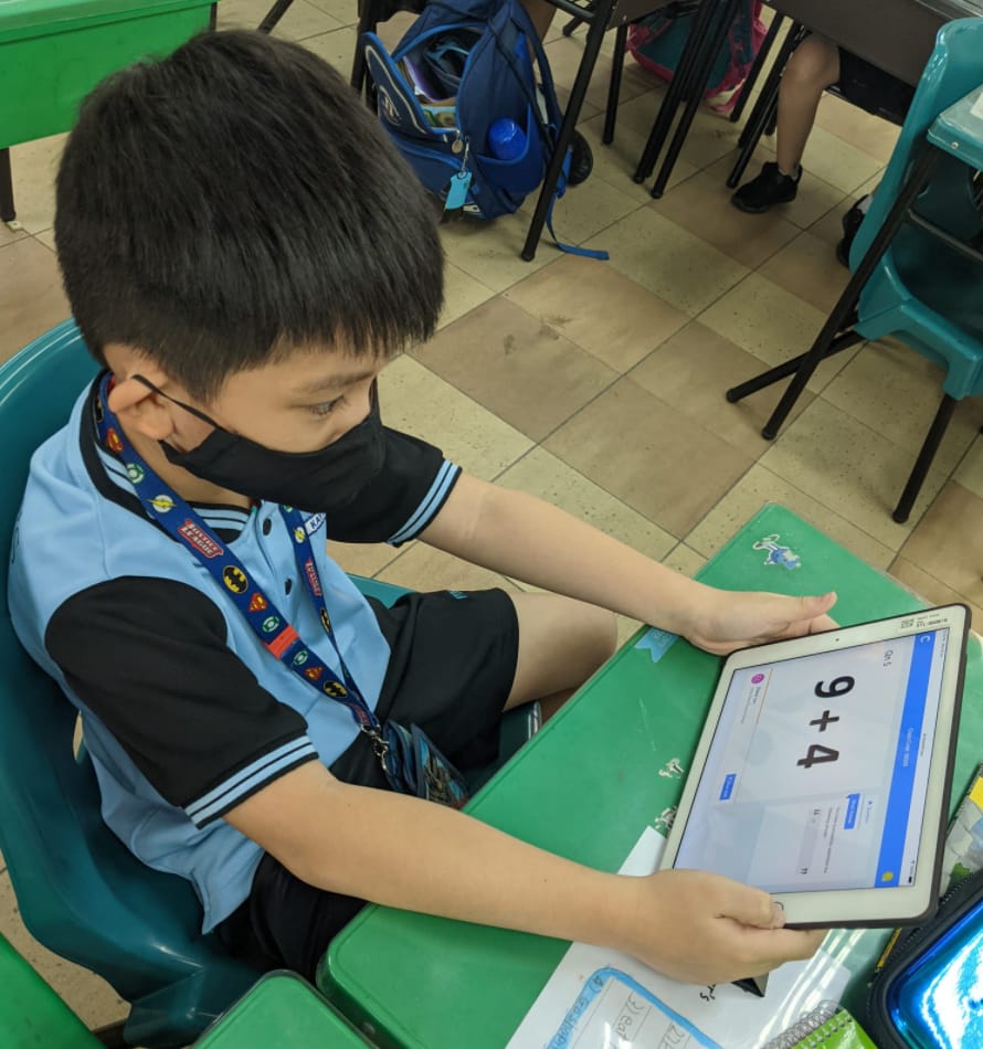 A young boy wearing a black mask sits at a green desk, holding a tablet showing the math problem 9+4.