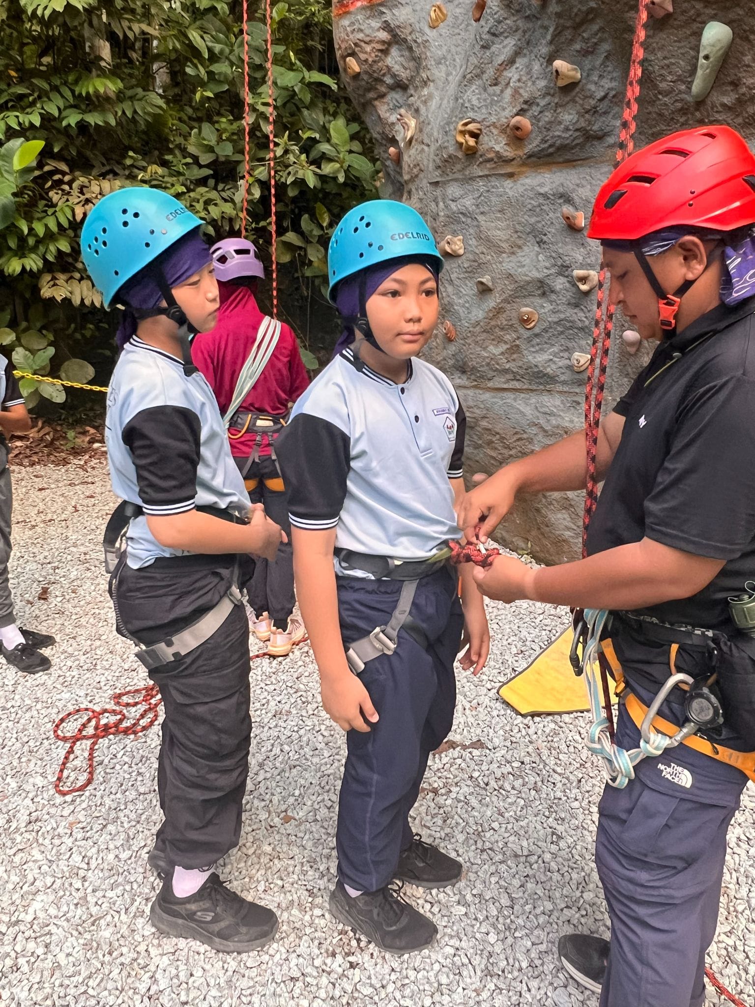 An instructor in a red helmet adjusts harness ropes for two children in blue helmets near a climbing wall.